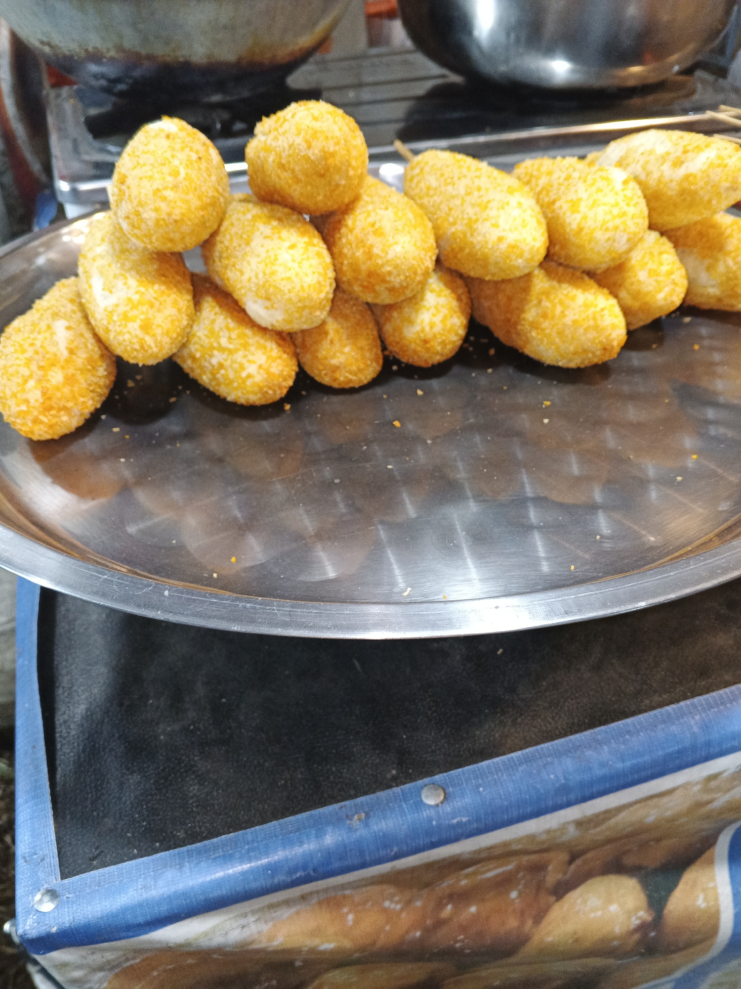 A close-up, high-angle shot focuses on a metal platter filled with numerous golden-brown, oblong-shaped snacks that appear to be coated in breadcrumbs or a similar textured coating. The snacks are arranged in a dense cluster. The platter rests on a dark surface, possibly a table or counter, with a blurry blue and white striped material visible in the lower portion of the frame. In the background, a portion of a stainless steel pot or large cooking vessel and a cooking stove are visible, suggesting a food preparation or serving environment. The lighting is uneven, creating some shadows and highlights on the snacks and platter. The image was taken in the city of Sukabumi, Indonesia.