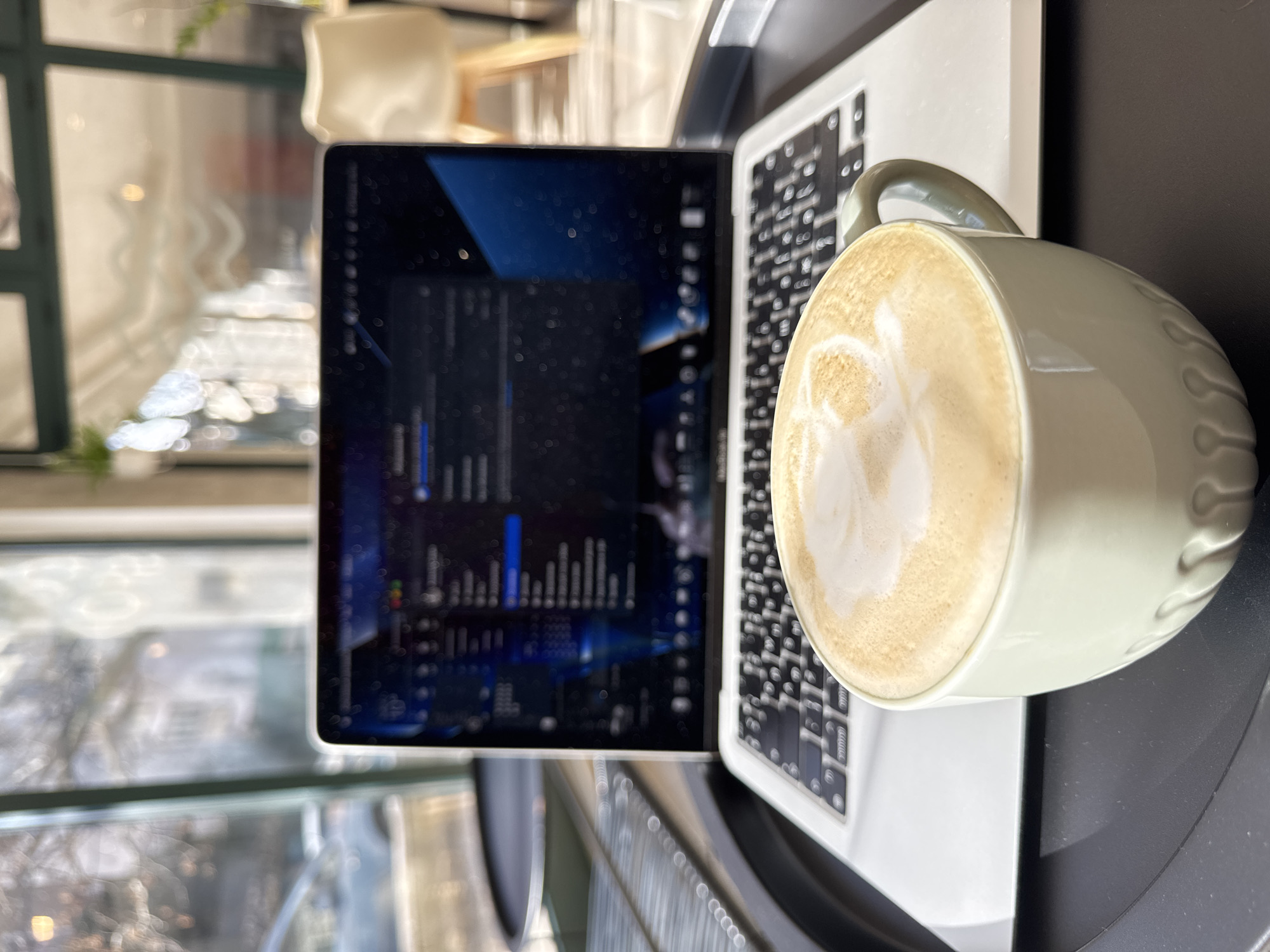 The image depicts an indoor scene within a commercial establishment, likely a cafe, located in an urban area of Budva, Montenegro. In the immediate foreground, a light sage-green ceramic mug, featuring vertical fluting on its lower half, is positioned centrally. The mug contains a latte or cappuccino, identifiable by the foamy milk art on its surface. This mug rests on a dark, circular tray or tabletop.
Behind the mug, a silver laptop, open and active, occupies the midground. Its screen displays a dark user interface with multiple application icons and window elements, including a prominent blue graphical element on the right side of the display. The laptop's keyboard is visible beneath the screen, showing a QWERTY layout.
Through large windows in the background, an out-of-focus outdoor urban environment is visible. To the left, a light-colored vehicle is partially discernible on what appears to be a street. Bare tree branches are also visible through the windows, suggesting a cold season. To the right, blurred interior elements include part of a light-colored chair with wooden legs and what appears to be tiled flooring, consistent with a cafe setting. No individuals are depicted in the frame. The scene is illuminated by natural light entering through the windows, causing some reflections on the laptop screen.