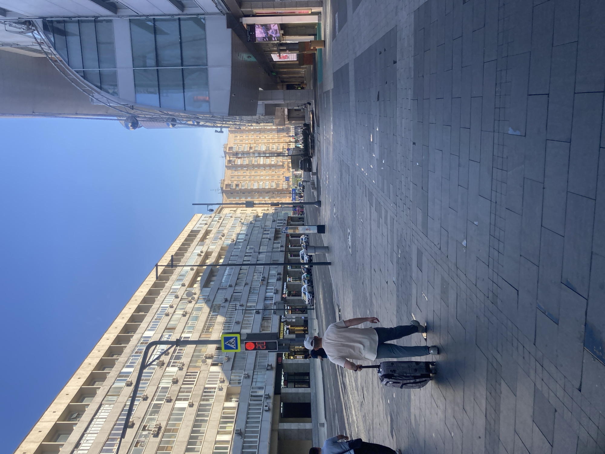 The image depicts a street scene in Moscow, Russia, with a focus on the pedestrian walkway in the foreground and buildings lining the street. In the foreground, a man wearing a light-colored shirt and a baseball cap is seen walking away from the camera, pulling a patterned suitcase on wheels. Another person, partially visible on the left, is walking in the same direction.
The walkway is paved with large, dark gray tiles, arranged in a somewhat irregular pattern. The street itself, which runs parallel to the walkway, is lined with cars. Traffic lights are visible, one showing a red pedestrian signal with a countdown timer.
Dominating the background are several multi-story buildings. On the left, a long, light-colored apartment building with numerous windows and balconies stretches across the frame. On the right, a modern building with a curved glass facade and metallic accents rises. Further in the distance, more apartment buildings, one with a distinctive light brown color, can be seen under a clear blue sky. The overall impression is an urban environment during the daytime, likely a commercial or residential area of the city.