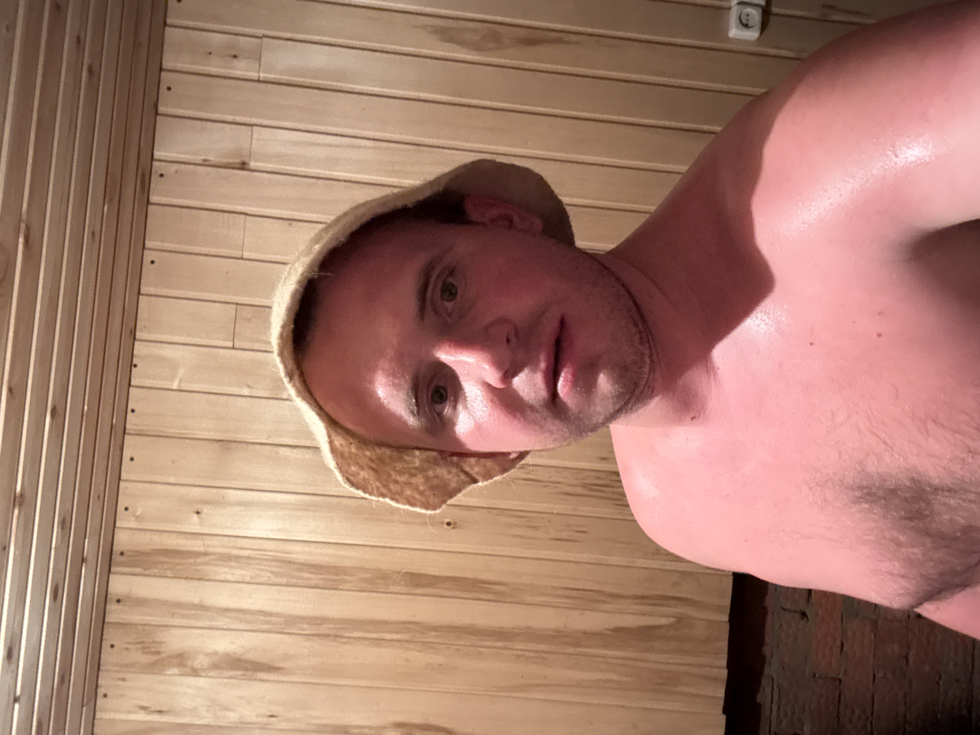 A shirtless man wearing a felt hat is indoors, likely in a sauna, due to the wooden paneling walls. His skin is red and glistening with sweat. The location details are not discernible within the image.