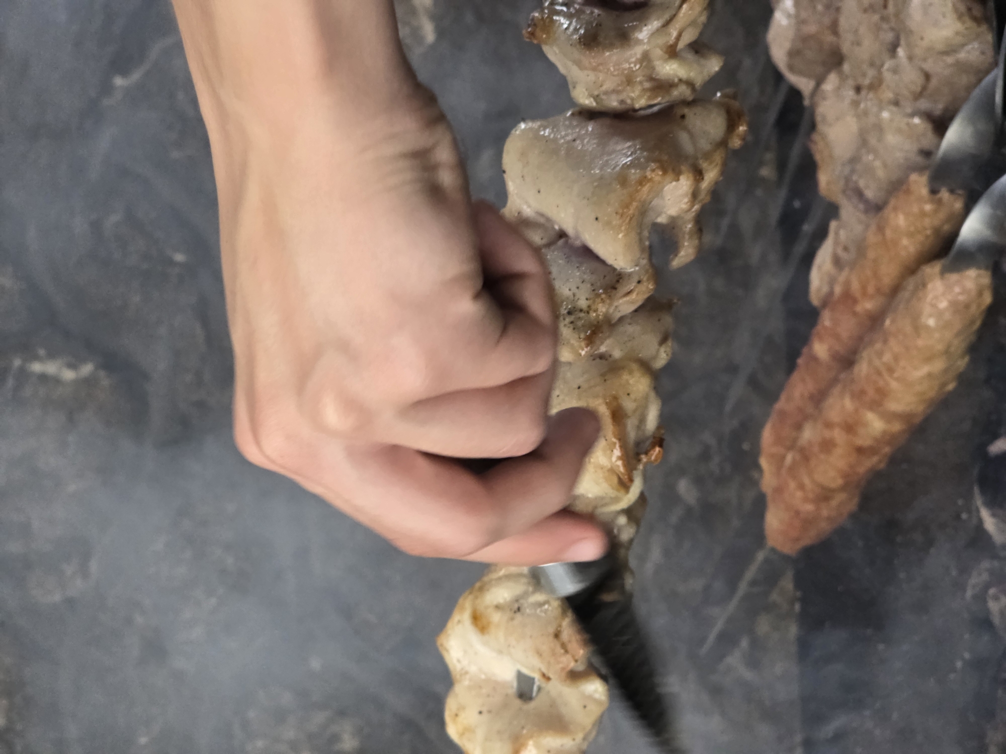 A close-up shot shows a hand manipulating meat on skewers over a dark, smoky background. The hand, with its pale skin, is positioned on the left side of the frame, fingers gently touching pieces of cooked meat threaded onto a metal skewer. The meat appears to be in various stages of cooking, with some pieces displaying a light brown char. The skewers themselves are visible, some holding chunks of meat and others holding what look like ground meat sausages. The smoky atmosphere suggests an outdoor cooking environment, possibly a barbecue or grill. The lighting is dim, indicating it might be evening or a shaded area. The overall scene evokes the preparation of a meal, likely a traditional dish involving grilled meat. The location context of Pidhorodne, Ukraine, hints at a possible cultural or culinary setting related to that region. There is no visible text in the image.