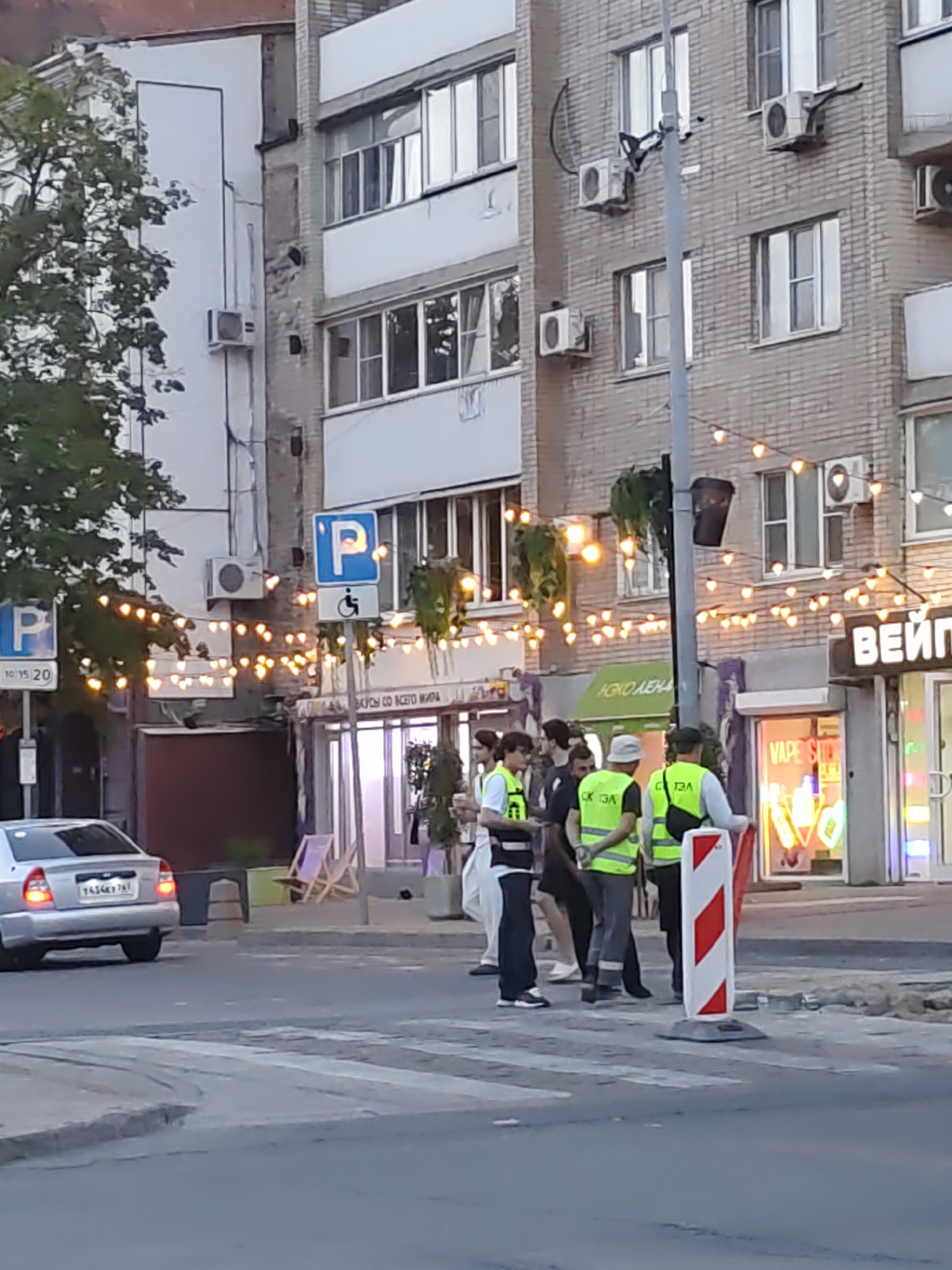 The image depicts a street scene in Rostov-on-Don, Russia, focusing on a crosswalk area adjacent to a building with multiple windows and air conditioning units. Several individuals are visible, some wearing high-visibility vests, suggesting a work or safety detail. One individual in the foreground holds a red flag, possibly indicating a temporary traffic control situation. A silver car is stopped at the crosswalk. Decorative string lights are hung across the street, illuminating the area. The building facade is made of brick, and there are signs and shop fronts visible, including one with "VAPE SHOP" lit in neon. The time appears to be late afternoon or early evening, judging by the lighting.