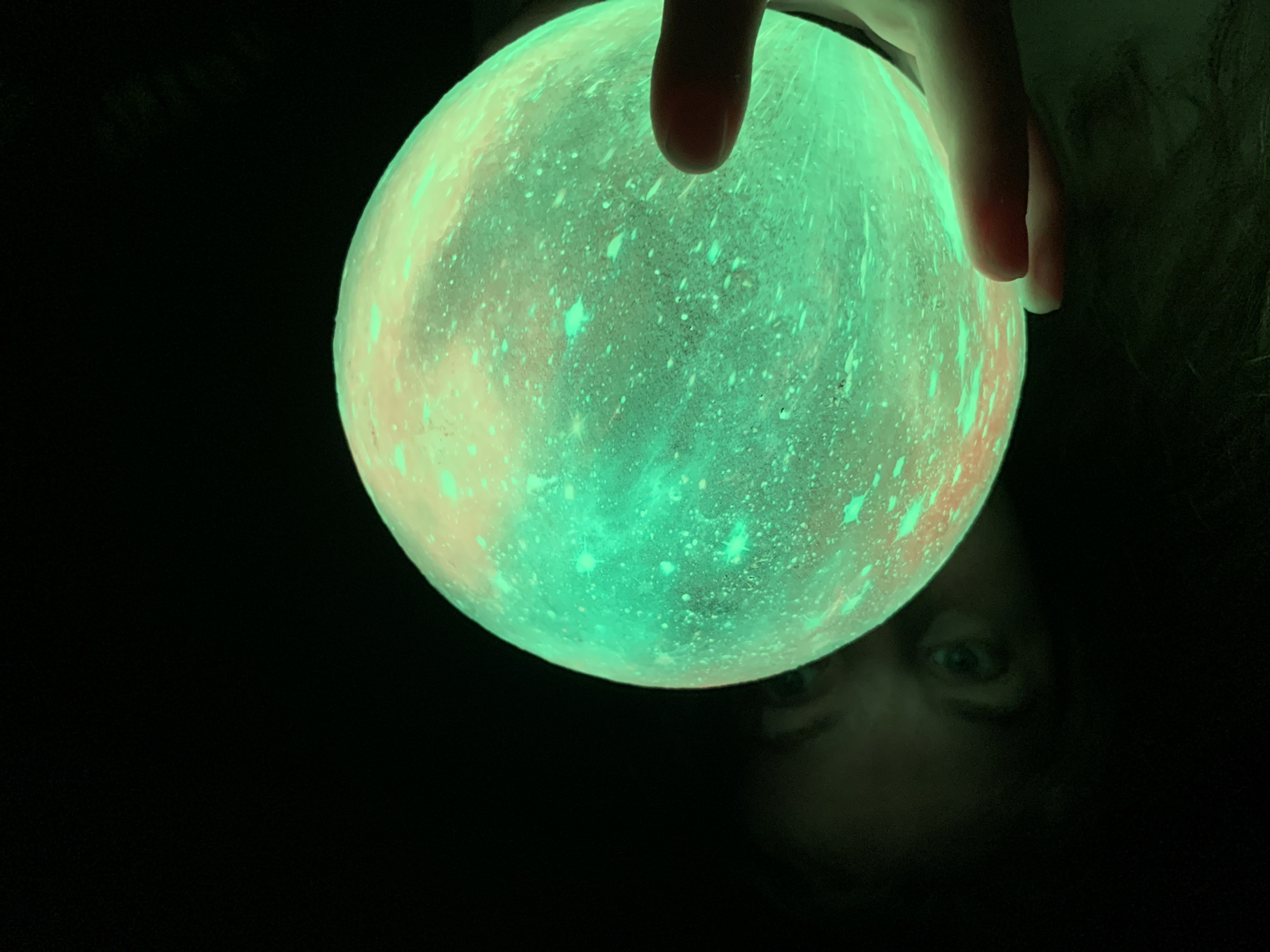 A glowing spherical object, emitting vibrant green light with yellow-orange accents and a textured, crater-like surface, occupies the central and right portions of the frame. A human hand, with fingers curled, grasps the lower right side of the sphere. In the darkened background on the left, two human eyes are visible, reflecting the green light from the object. The surrounding environment is predominantly dark, suggesting a low-light setting. This scene is located in Chaikovsky, Russia.