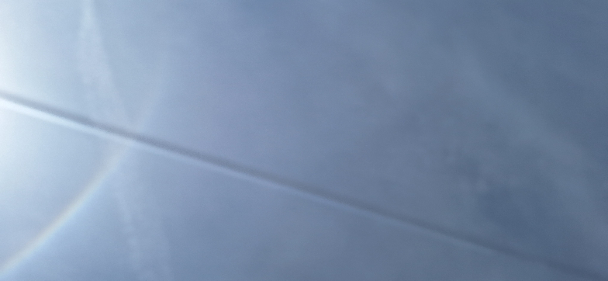 The image shows a blurred view of a pale blue sky with a horizontal line bisecting the frame diagonally from the lower left to the upper right. A faint rainbow or prismatic effect appears in the lower left corner, suggesting a possible light refraction phenomenon. The out-of-focus nature of the image obscures any finer details about the sky or potential subjects within it. There is no information in the image to determine a location in Blagoevgrad, Bulgaria.