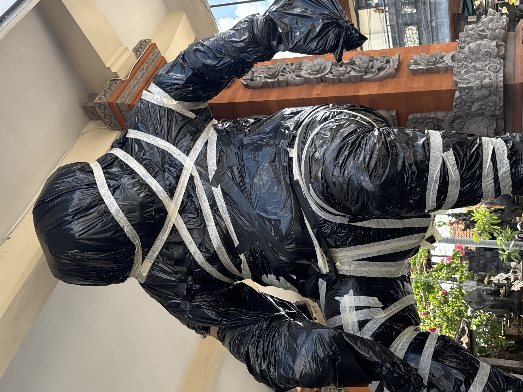 A full body shot from a low angle shows a large, human-like figure wrapped in black plastic sheeting and secured with white tape. The figure is positioned outdoors, with traditional Balinese architecture and lush greenery visible in the background. The architecture features intricate stone carvings on red brick pillars, and flowering plants with pink blossoms add color to the scene. The sky above is partly cloudy. The figure appears to be standing or in a dynamic pose, with one leg forward. The lighting suggests it is daytime. The location is identified as Ubud District, Indonesia.