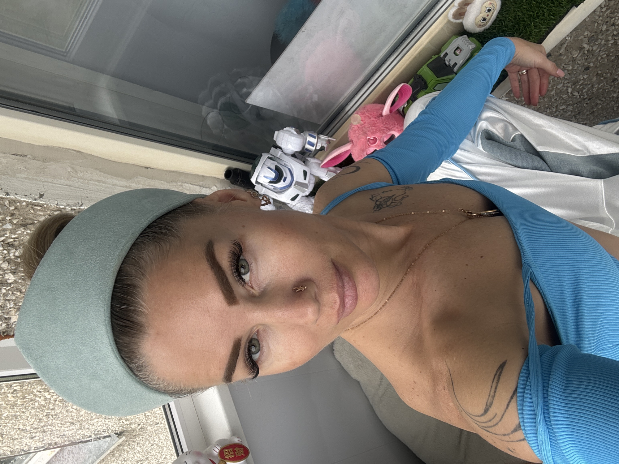 A woman wearing a light blue headband and a matching blue top is positioned indoors with a partial view of a window and some toys. The background environment reveals a window sill with a collection of items including a white robot toy, a pink plush rabbit, a green toy vehicle, a sheep-shaped plush toy, and faux grass. The location and city are not discernible from the image content.