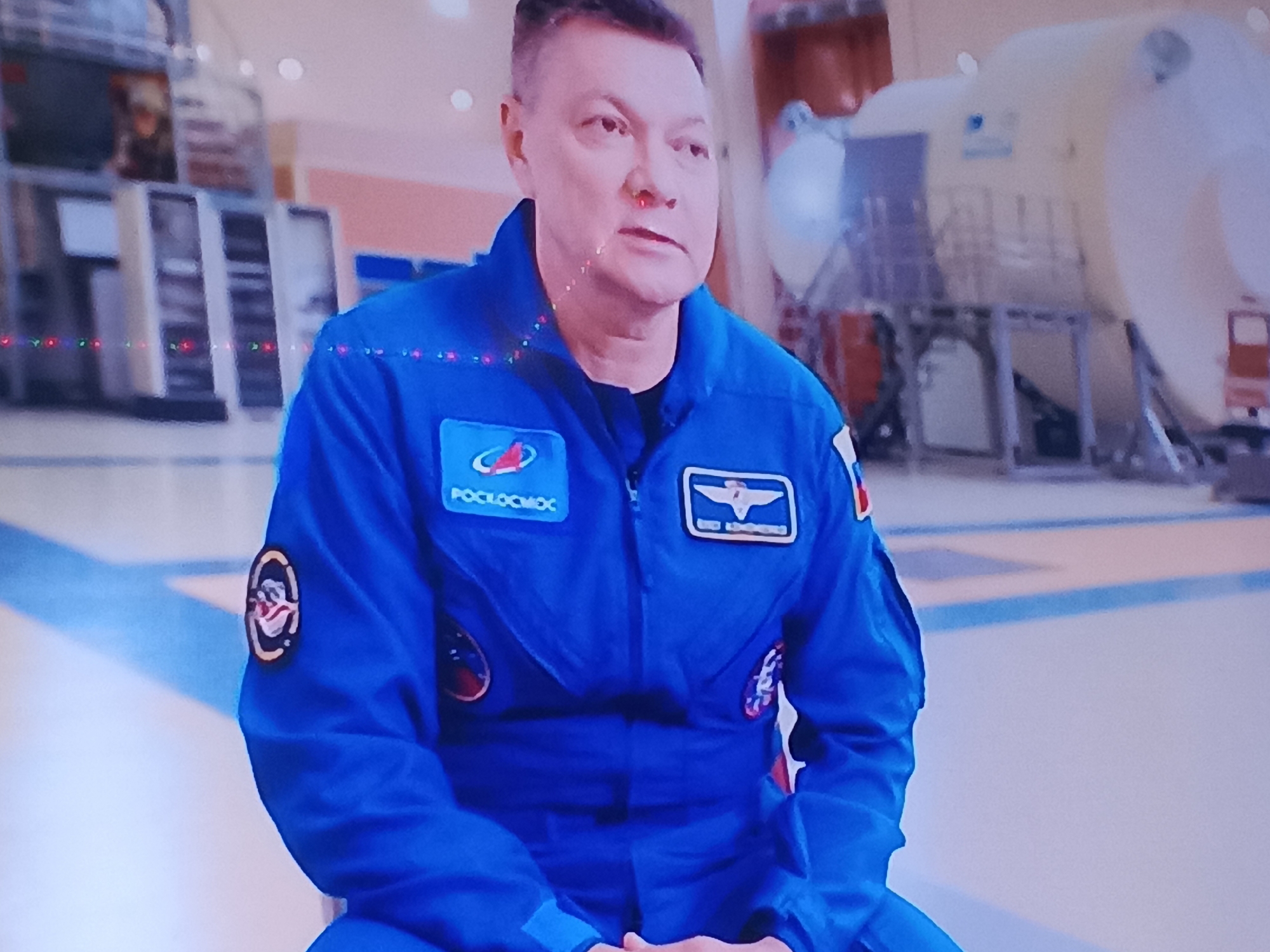 The image shows a man in a blue flight suit, possibly an astronaut, sitting down. The flight suit has patches on it, including one that says "РОСКОСМОС". In the background, there is some sort of module or spacecraft-like structure. The lighting suggests an indoor environment, possibly a museum or training facility. The man's gaze is directed to his right. There are no visible signs or indications that this scene is located in the city of Biysk, Russia.