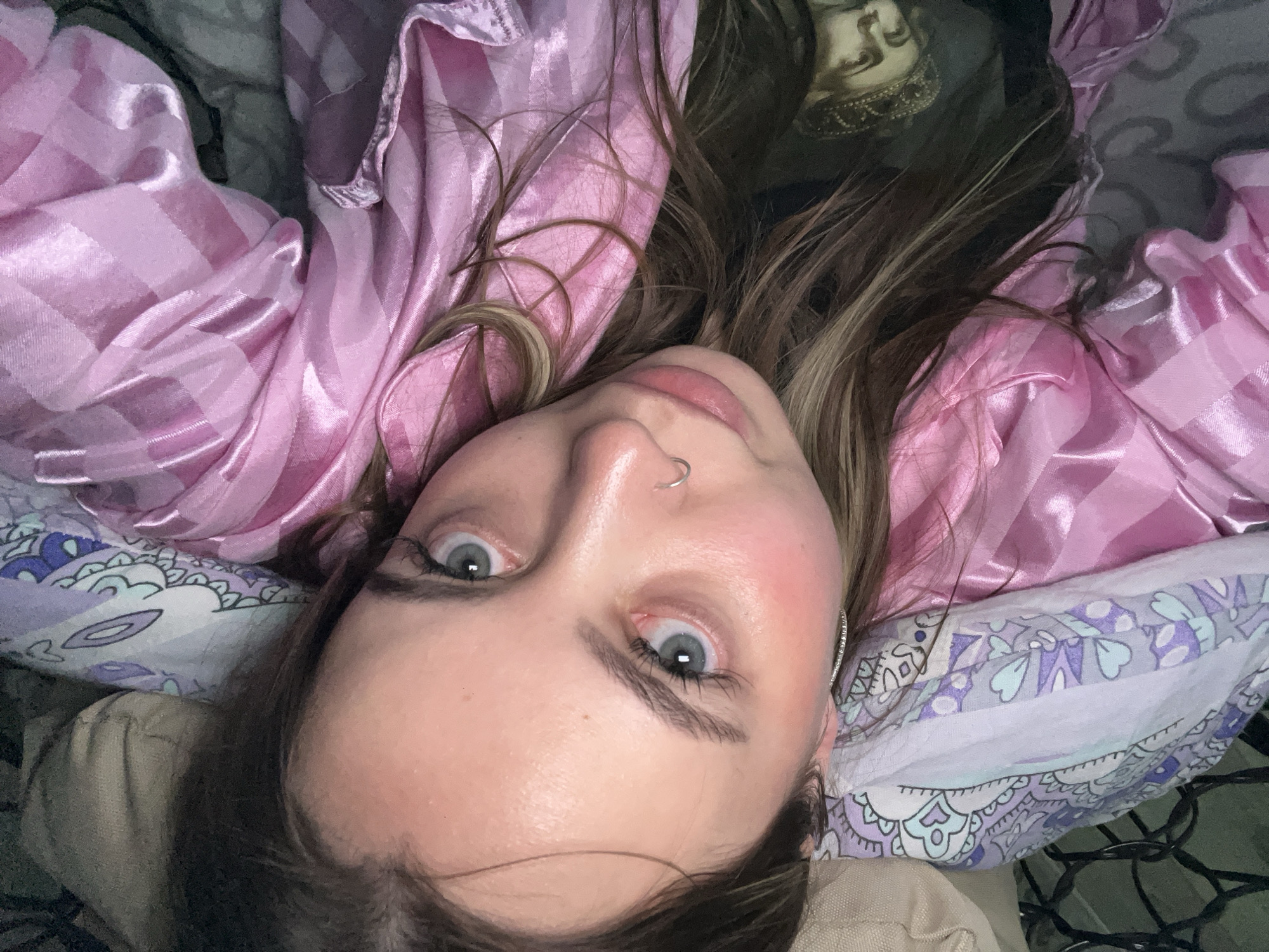 A young woman is lying on a bed. She is wearing a pink shirt and has light eyes. Her head rests on a patterned pillow. An object with an image of a face is visible in the upper right corner. The scene's location is not discernible in the image.
