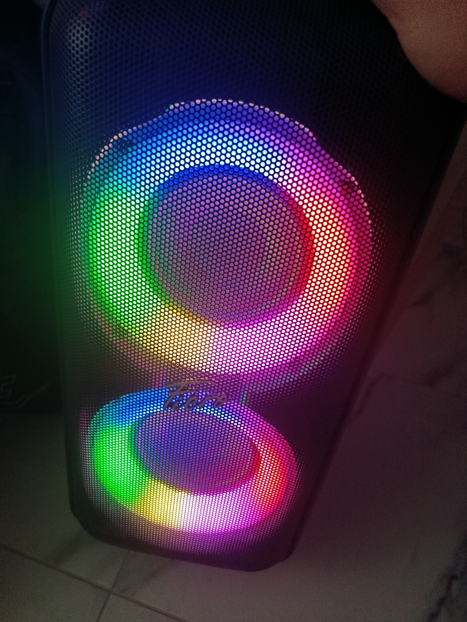 A close-up, low-angle shot shows the front of a tall, black speaker with two illuminated circular grilles. The grilles are lit with a rainbow of colors, including green, yellow, orange, red, pink, and blue. The speaker appears to be standing on a tiled floor. The background is dark and out of focus, with a hint of a hand visible in the upper right corner. The image is likely taken indoors, possibly in a home or a studio. There is no visible information in the image that would place it in Vyselki, Russia.