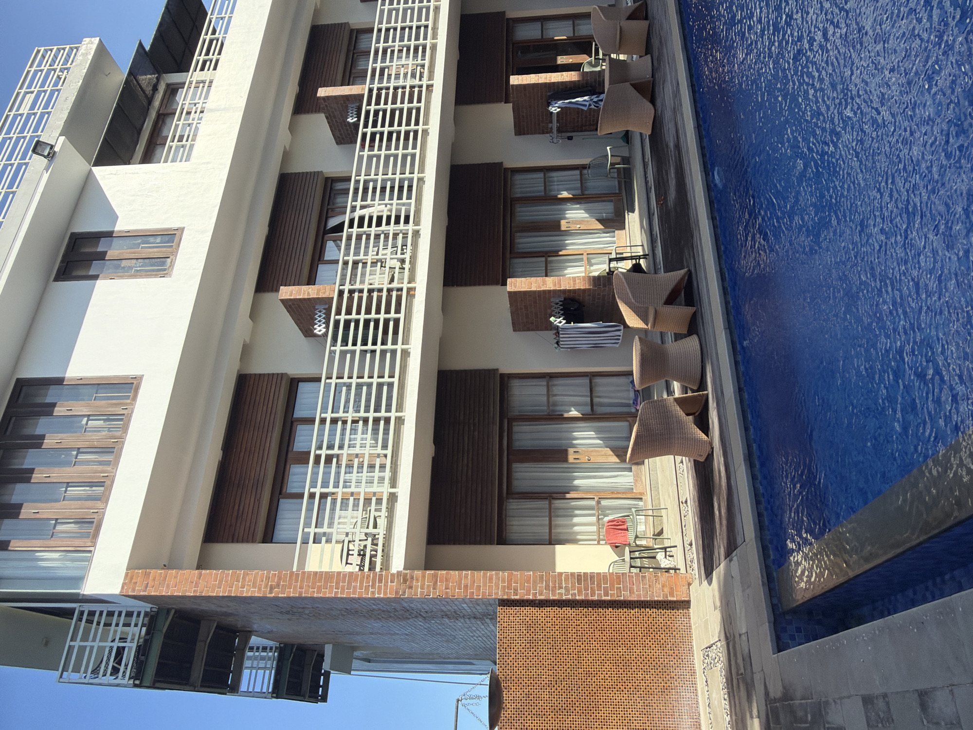 The image depicts a hotel in Kuta Selatan, Indonesia, with a swimming pool in the foreground. The hotel is a multi-story building with a white facade and wooden-framed windows. Red brick accents are visible on the exterior walls and around some of the windows. Balconies with white railings extend from the upper floors.
In front of the hotel, a blue tiled swimming pool occupies the lower portion of the frame. The pool has a clean, bright blue color and appears to be well-maintained. The area surrounding the pool is paved with stone tiles.
On the patio area adjacent to the pool, several wicker chairs and small tables are arranged. These appear to be outdoor furniture for guests to relax. A red towel is draped over one of the chairs. Various items, including towels and an umbrella, are hung on a clothesline near one of the ground-floor rooms. The overall scene suggests a tranquil and inviting resort environment. The sky above is a clear, bright blue, indicating a sunny day.