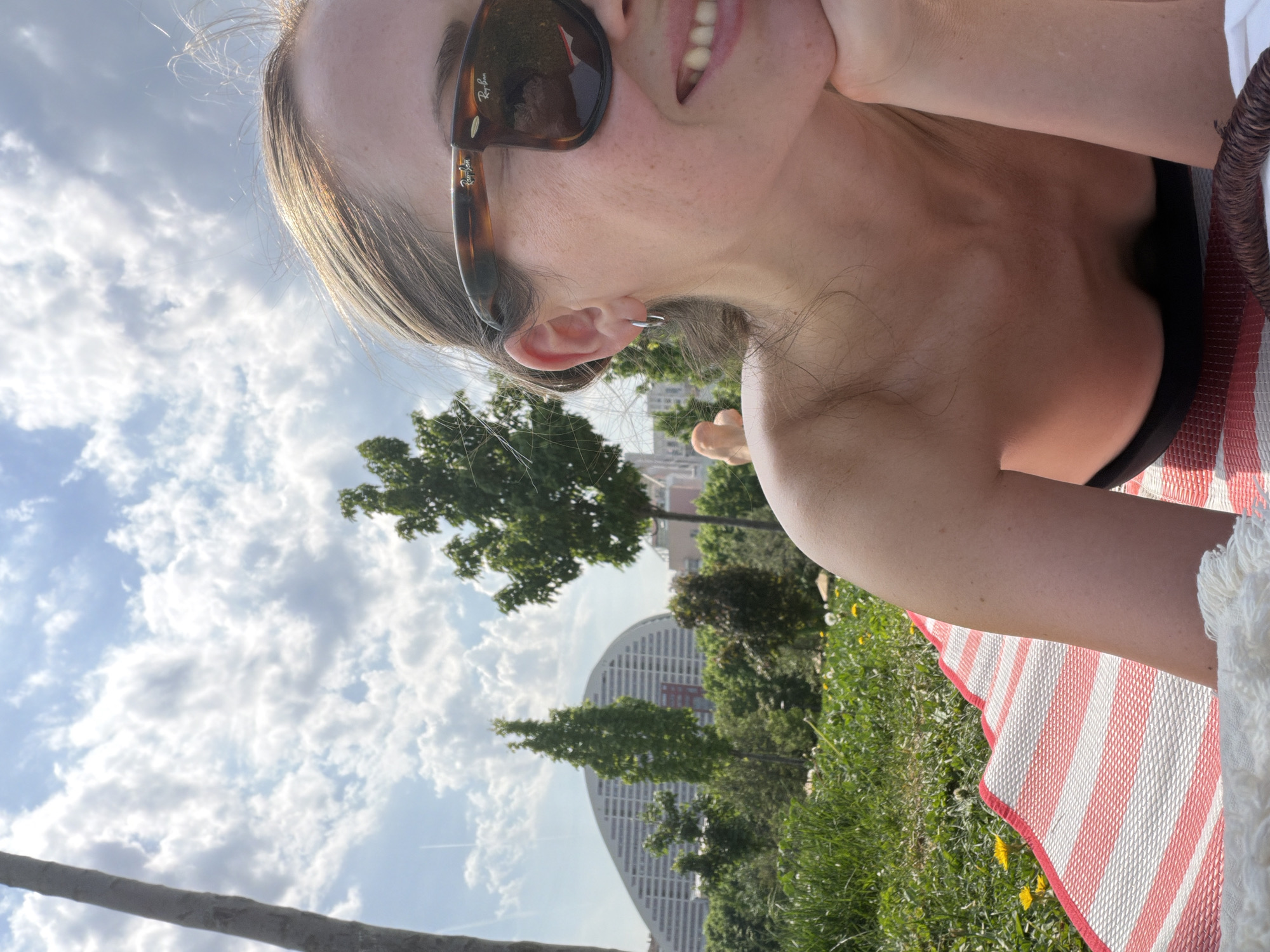 A woman wearing Ray-Ban sunglasses and a black top is lying on a red and white striped mat in a grassy area. Her right hand is supporting her head. In the background, there is a tree and a tall, curved building, similar in architecture to "Capital City" in Moscow, which is located in Khimki, Russia. The sky is cloudy.