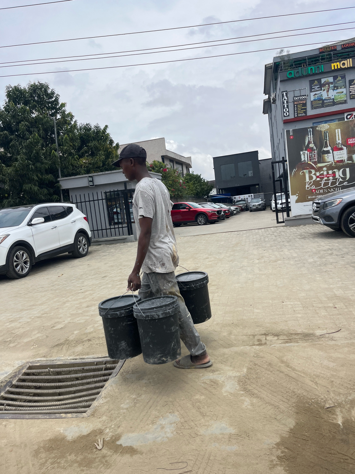 A person walks carrying two large black buckets in Ado, Nigeria. The person is wearing a dirty white t-shirt, grey pants, a black baseball cap, and sandals. The buckets appear to be heavy and are being carried by their handles. The ground is dusty and there is a metal grate in the foreground. In the background, there is a white SUV, a fence, trees, and a building with signs that say "Adunni Mall" and "Bellies Lounge". The sky is cloudy.