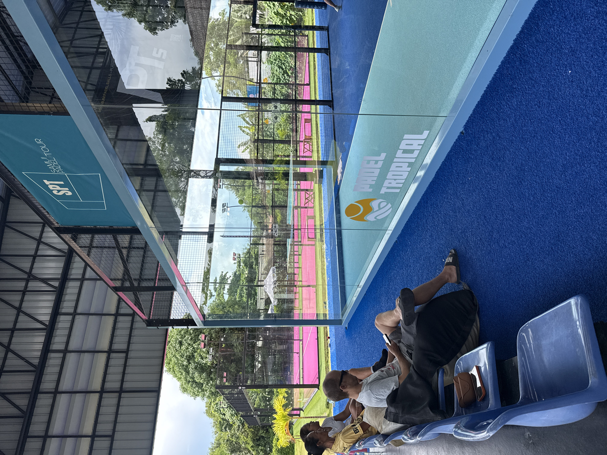 The image depicts an outdoor padel tennis court facility in Ban Bang Rak, Thailand. In the foreground, several individuals are seated on blue plastic chairs, some of whom are looking at their phones. The seating area is adjacent to a glass-enclosed padel court with blue and light blue carpeting. The court itself features pink playing surfaces and is surrounded by black fencing. A large overhead roof structure with metal supports covers part of the facility. In the background, lush green trees and vegetation are visible, along with additional padel courts and outdoor lighting. A teal-colored banner with the SPT Samui Padel Tour logo is mounted on the roof structure. Reflections in the glass court reveal more of the surrounding environment and parts of the court's interior.