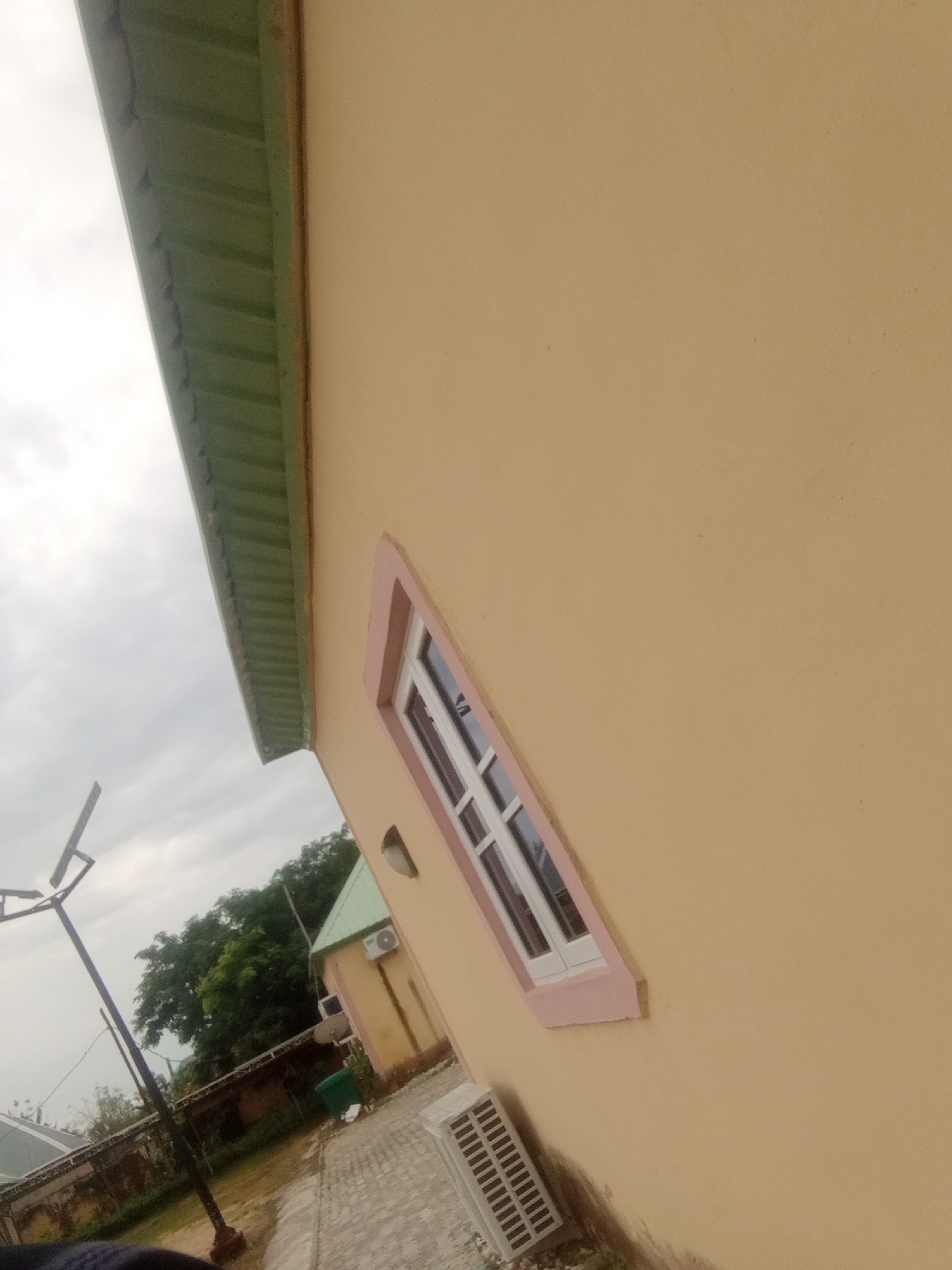 The image shows an outdoor scene in Jalingo, Nigeria, featuring the exterior of a building with light brown walls. A green-tiled roof is visible along the top edge of the frame, contrasting with the wall's color. A small, angled window with a white frame and pink trim is set into the wall. Below the window, an air conditioning unit is mounted on the wall.
To the left of the building, a tall, dark pole with what appears to be solar panels or a light fixture is positioned. Further in the background, another building with a green roof and an air conditioning unit can be seen, along with lush green trees. The ground in front of the buildings is paved with interlocking bricks. A dark-colored trash bin is situated near the second building. The sky is overcast, suggesting a cloudy day. The image is taken from a low angle, looking upwards towards the building and sky. There is no visible text in the image. The scene appears to be part of a residential or commercial property in a tropical environment.