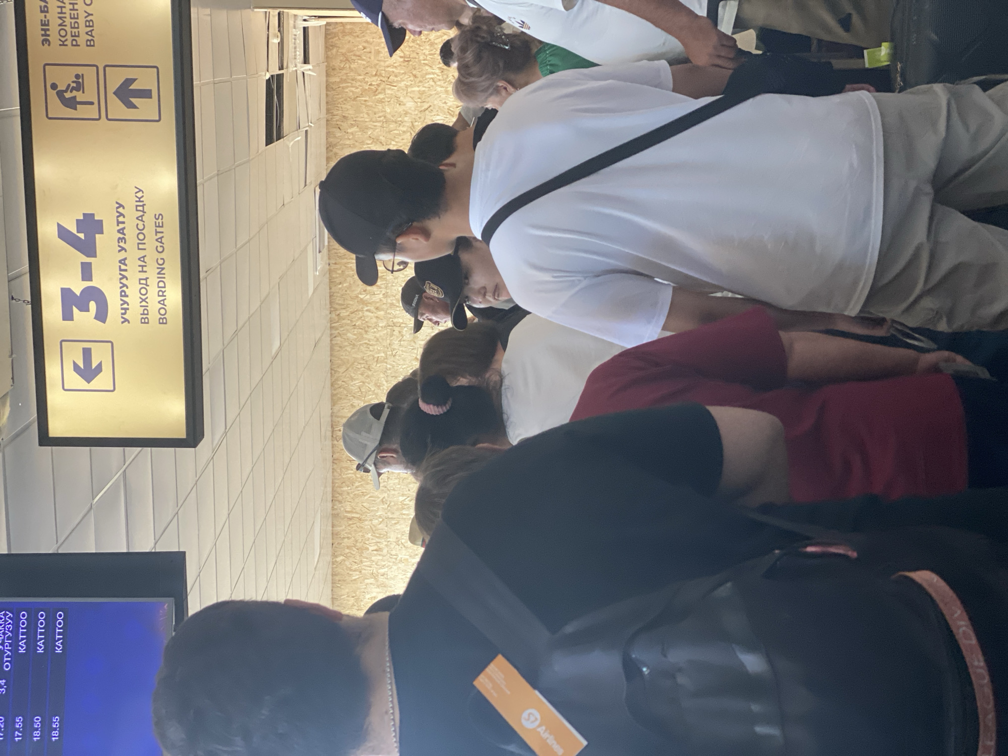 The image captures a scene inside what appears to be an airport terminal in Bishkek, Kyrgyzstan. In the foreground, several people are visible from the back, suggesting they are queuing or waiting. One individual in a black t-shirt has a black backpack with an orange tag that reads "ST Airlines". To the right, a person in a white t-shirt and tan shorts is also seen from behind. Above them, a large sign with purple text indicates "3-4" and "BOARDING GATES" in English, alongside directional arrows. Another sign to the right features a symbol of a person tending to a baby and an upward arrow, suggesting a baby care room and a different direction. In the background, a departure board displays flight information with times and destinations, including "KATTOO". The walls have a light-colored wood paneling texture. The overall impression is a busy, functional airport environment.