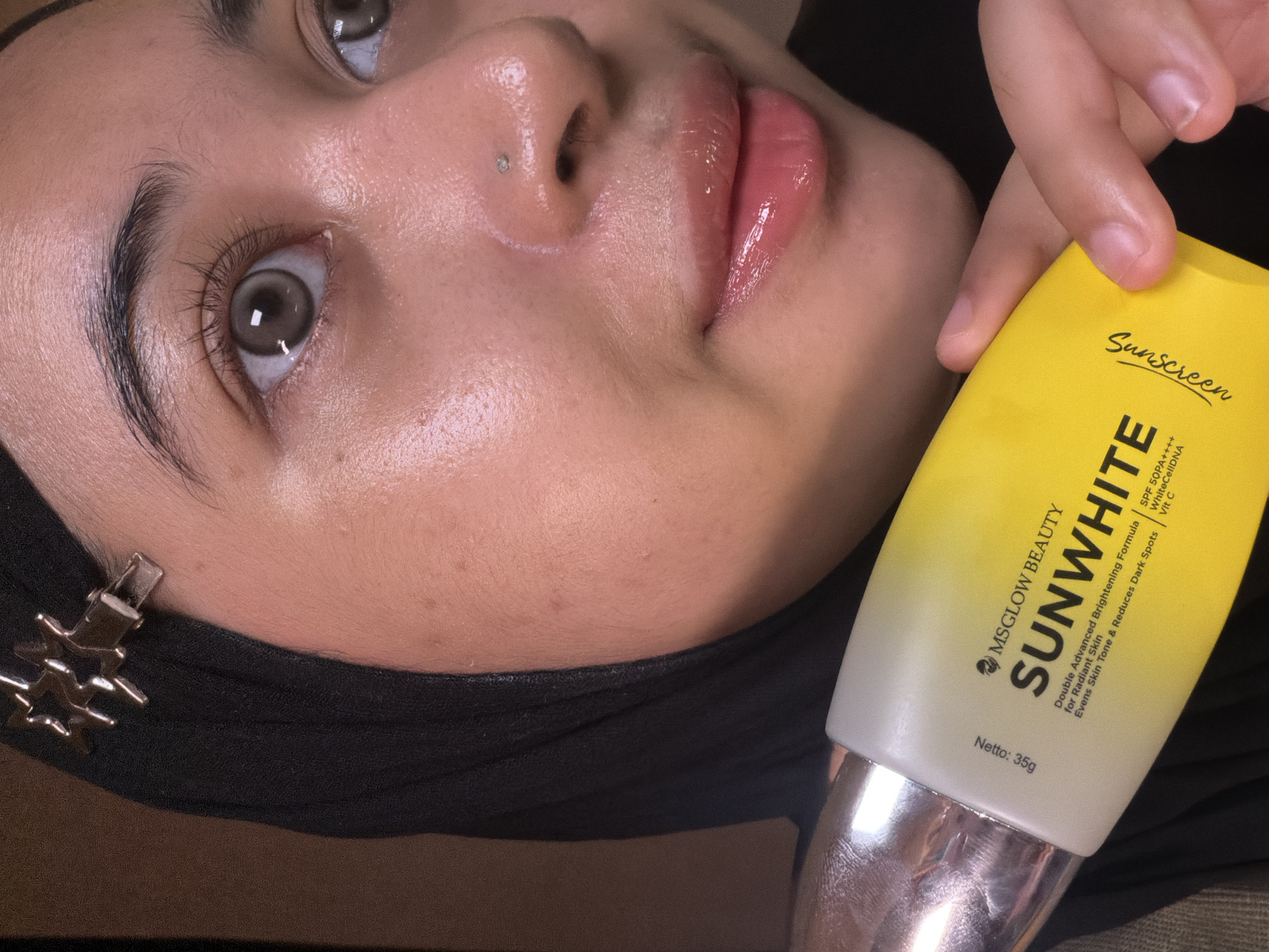 An upward-facing close-up shot captures a person holding a tube of MSGLOW BEAUTY SUNWHITE Sunscreen. The person's face is partially visible, featuring a nose piercing with a small stud, glossy pink lips, and visible eyes that appear to have light-colored contact lenses. Dark, defined eyebrows frame the eyes, and the skin exhibits a dewy, luminous finish. A dark head covering, possibly a hijab, is worn, secured by a silver-colored hair clip visible on the left side of the frame.
The sunscreen product, held in the person's right hand, is a yellow and white tube with silver accents at the bottom. The label prominently displays "MSGLOW BEAUTY" and "SUNWHITE" along with "Sunscreen." Further text on the tube highlights its benefits: "Double Advanced Brightening Formula for Radiant Skin," "Evens Skin Tone & Reduces Dark Spots," "SPF 50PA++++," "WhiteCellDNA," and "Vit C." The net weight is indicated as "Netto: 35g."
The lighting appears to be soft, creating highlights on the person's skin and the glossy lips, suggesting an indoor setting. The image likely serves to showcase the skincare product, possibly for an advertisement or review. This scene takes place in East Jakarta, Indonesia.
