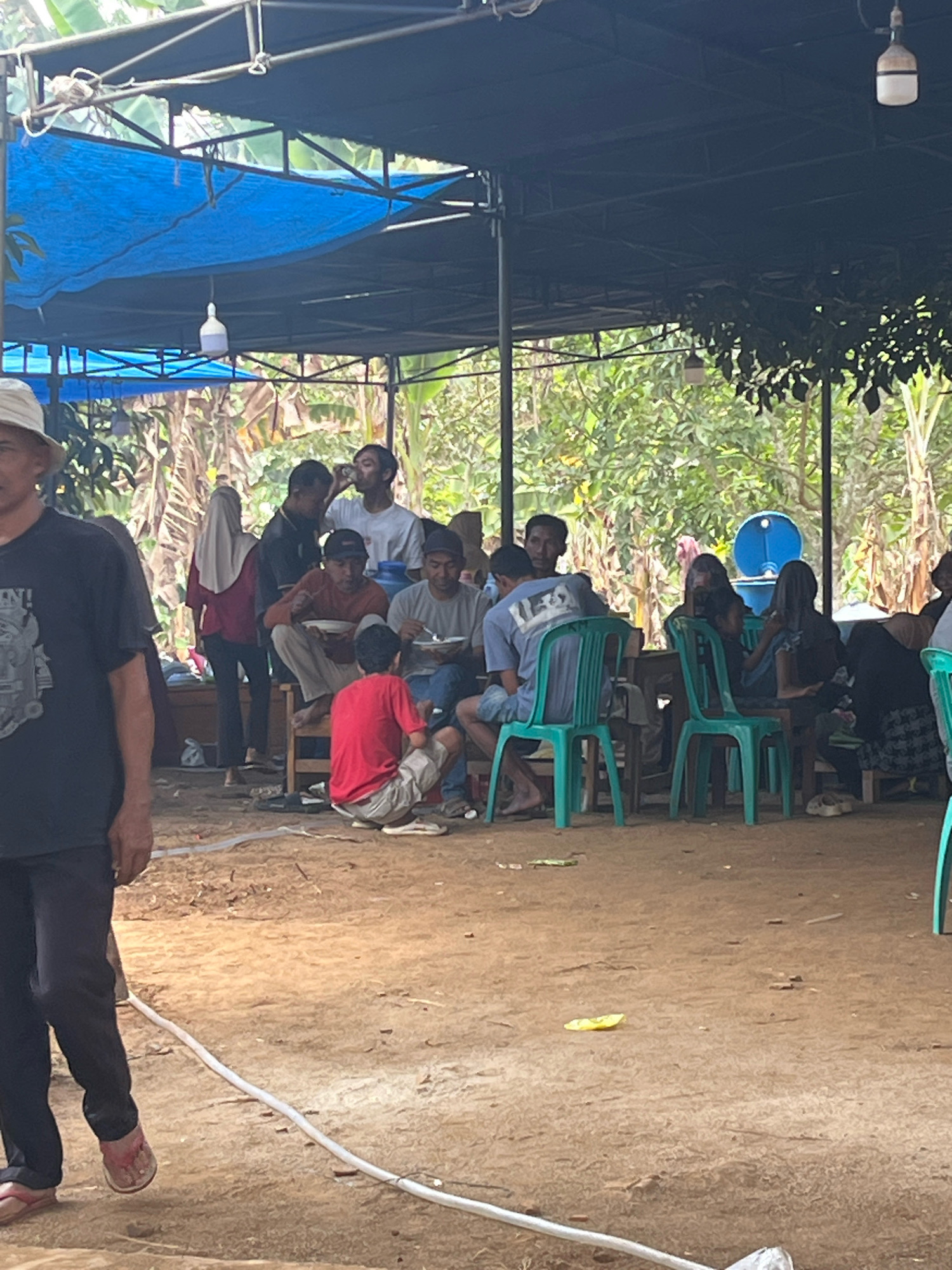 A group of people are gathered under a dark blue tarp, likely for an outdoor event or meal, in a dusty, outdoor setting. Some individuals are seated at tables, eating or drinking, while others stand or walk. The environment appears rural, with dense foliage visible in the background. The people are dressed casually, and the overall atmosphere seems relaxed. It is a scene from the city of Kebumen, Indonesia.