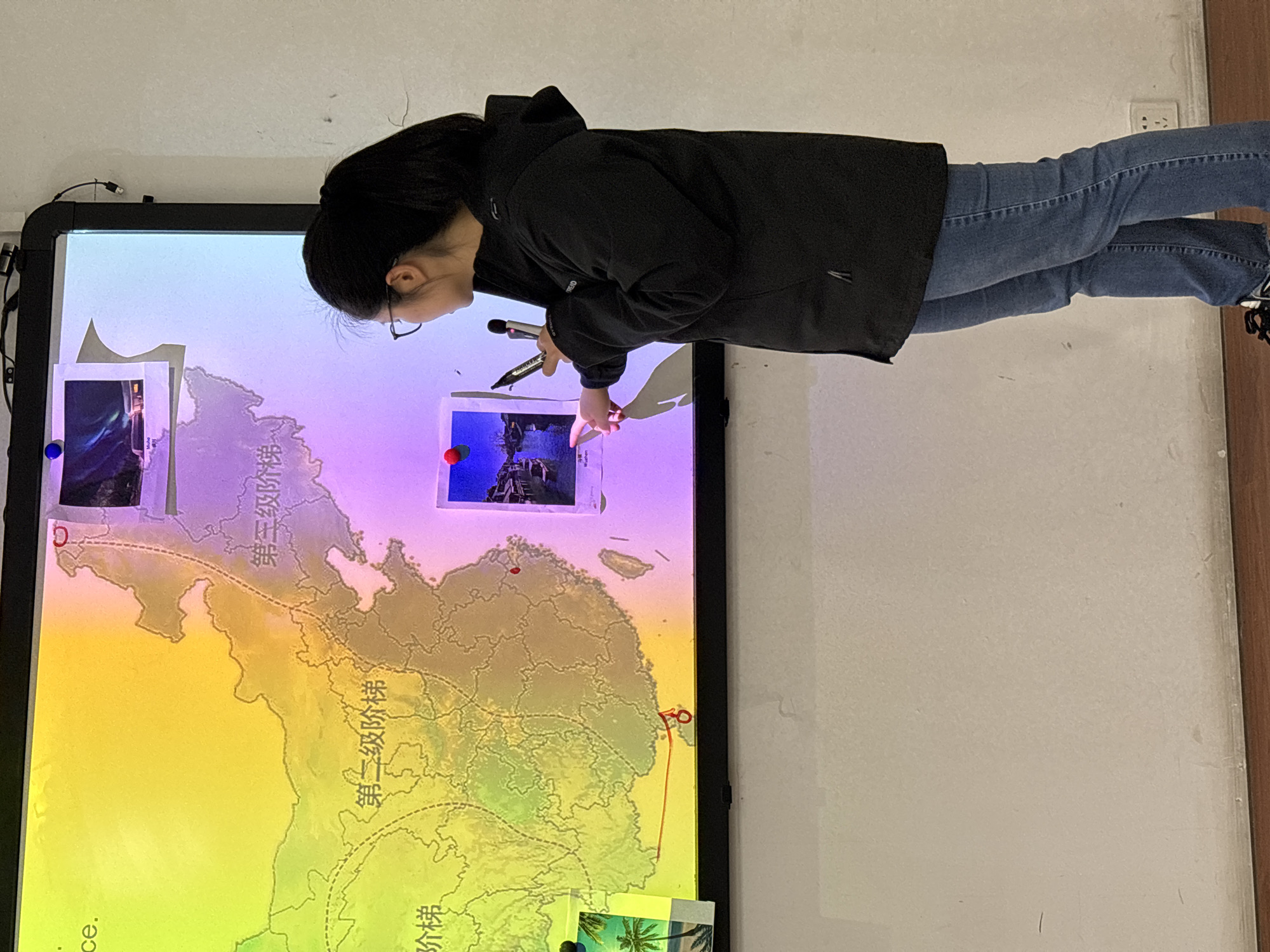 A person wearing glasses and a black jacket stands in front of a large interactive whiteboard, using a marker. The person has dark hair and is wearing blue jeans. The whiteboard displays a map of what appears to be the city of Jiangjin, China, and possibly surrounding areas. The map is overlaid with colorful gradients and marked with Chinese characters and images. The board has pictures tacked onto it with tacks. The scene is set indoors, likely in an educational or office environment with white walls.