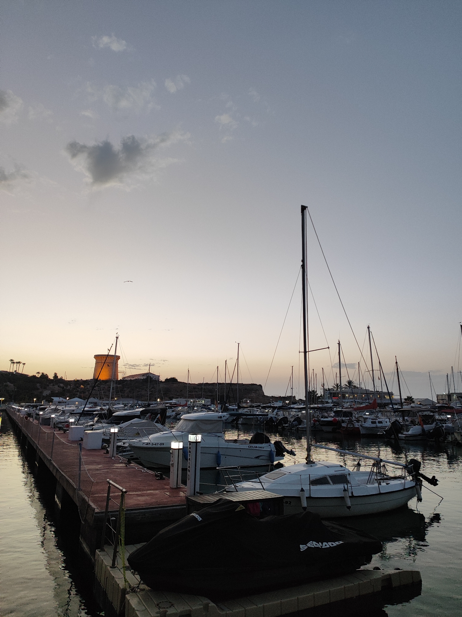 The image depicts a marina in El Campello, Spain, at dusk. A long, red-colored pier juts out into the water, lined with moored boats. Several yachts and smaller vessels are docked, some with covers over them. The water is calm and reflects the sky and the lights from the pier. In the background, a distinctive round, orange tower stands on a hill, illuminated by lights, with palm trees and other structures nearby. The sky is a soft gradient of pale blue and orange, with a few wispy clouds. A bird can be seen flying in the distance. The overall atmosphere is tranquil and serene.