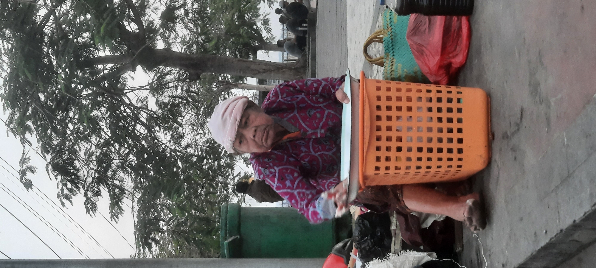An elderly woman wearing a light-colored headscarf and a dark patterned shirt is sitting on a concrete sidewalk in Kanigoro, Indonesia. She is leaning forward, her hands appearing to be engaged in an activity involving a white tray resting on an orange plastic basket. The basket is filled with white items. Behind the woman, a large green garbage bin is visible. Further in the background, several people are seated on the ground near a tree. The ground is a textured concrete surface. Several bags, including a green woven bag and a red plastic bag, are placed around the woman and the basket. The sky is overcast, and trees with dense foliage are present in the background, some with power lines running through them.