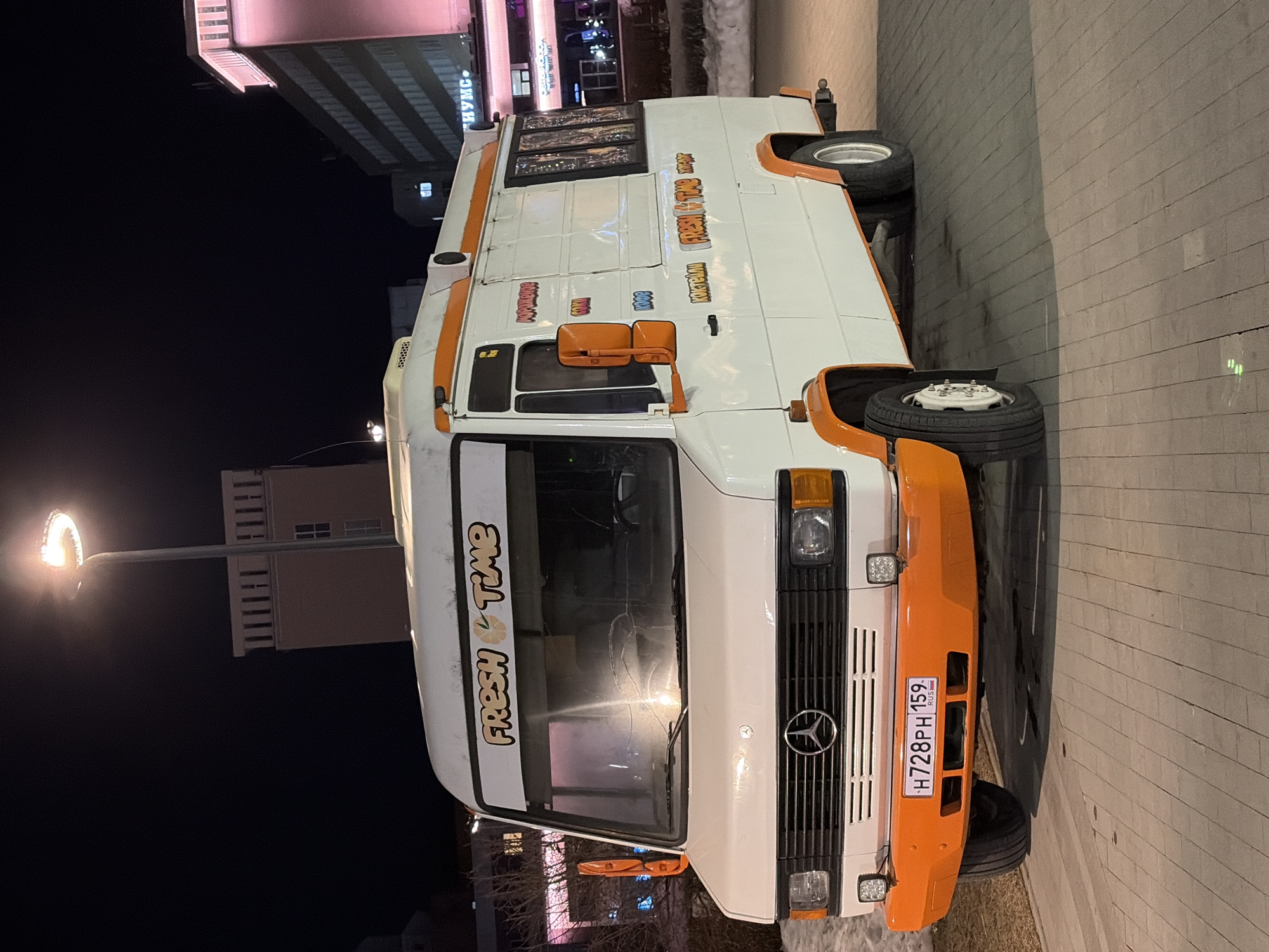 A white Mercedes-Benz van, converted into a food truck, is parked on a paved surface during nighttime in Perm, Russia. The vehicle features orange accents on its front bumper, wheel arch flares, and side mirrors, along with an orange stripe running along the roofline. The front windshield displays "FRESH TIME" branding and exhibits a distinct crack pattern. A Russian license plate, H728PH 159 RUS, is affixed to the front bumper, flanked by two square LED auxiliary lights. The right side of the van is visible, bearing additional "FRESH & TIME" branding and Russian text, likely advertising products such as "МОРОЖЕНОЕ" (Ice Cream), "СОКИ" (Juices), "КОФЕ" (Coffee), and "КОКТЕЙЛИ" (Cocktails). The van is illuminated by a prominent overhead streetlight situated to its rear-left. The surrounding area consists of light-colored rectangular pavers, with patches of snow visible on the ground. In the background, multiple buildings are discernible, including a tall, light-colored structure with intricate upper facade details and another building displaying neon pink illumination and partially visible Cyrillic text ("НУМС"). No individuals are present in the scene.