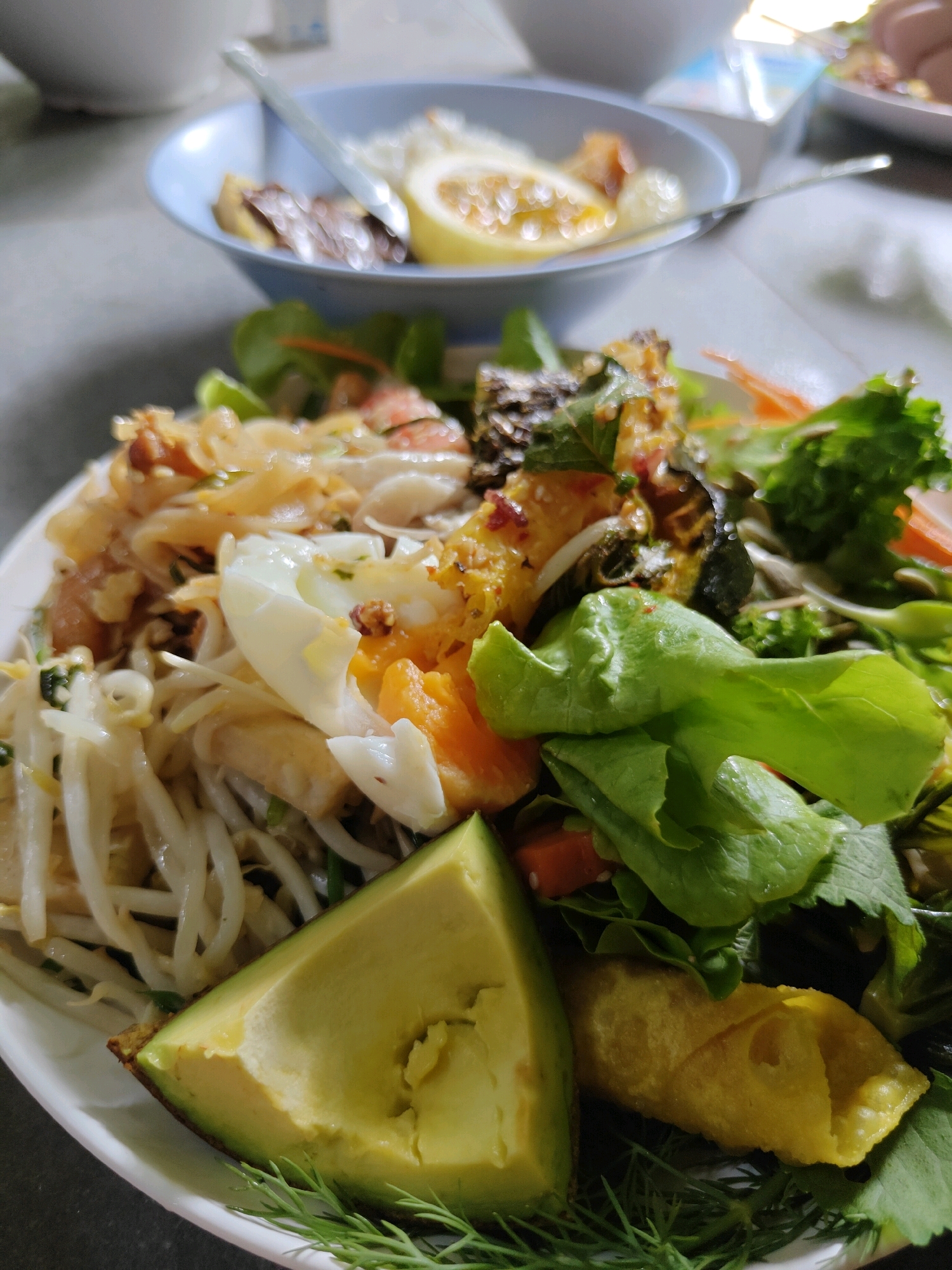 A close-up, overhead shot captures a vibrant plate of food, likely in a restaurant or eatery in Mu Si, Thailand. The dish appears to be a noodle salad, featuring a generous portion of white bean sprouts, shredded noodles, pieces of boiled egg, and what looks like shredded chicken or fish. Amongst these, bright green leafy vegetables and hints of orange carrot are visible, adding to the dish's visual appeal. A wedge of ripe avocado rests on the left side of the plate, its creamy texture contrasting with the other ingredients. A golden-brown, fried spring roll or similar pastry sits on the right, adding a textural element to the meal. In the blurred background, a blue bowl with some contents and a spoon are present, suggesting other dishes or accompaniments. The lighting suggests an indoor setting, possibly with natural light.