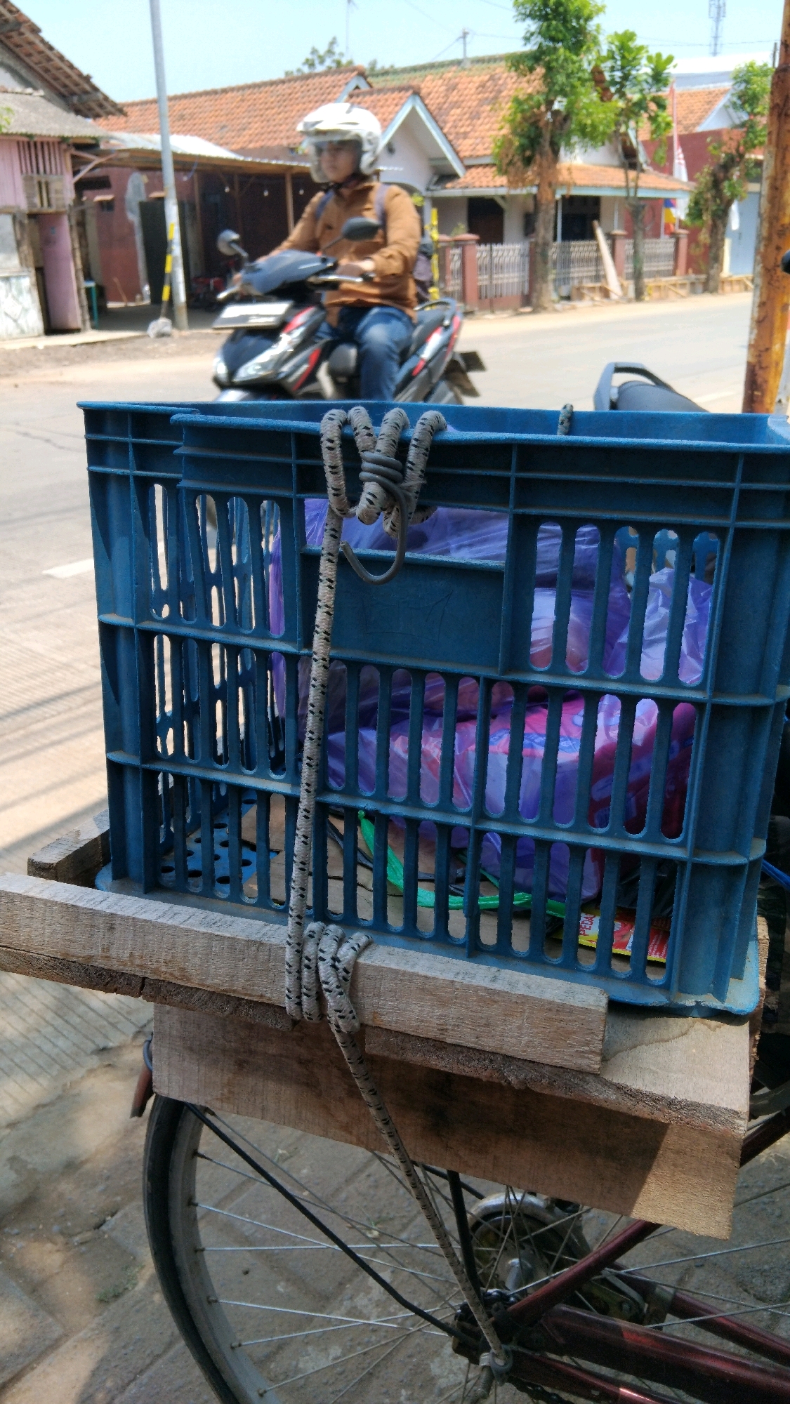In Batang, Indonesia, a person wearing a white helmet and an orange jacket rides a black motorcycle down a street lined with houses. In the foreground, a blue plastic crate is attached to the back of a bicycle, filled with items wrapped in purple plastic bags. A rope is used to secure the crate to the bicycle. The sky is clear and blue, indicating a sunny day.