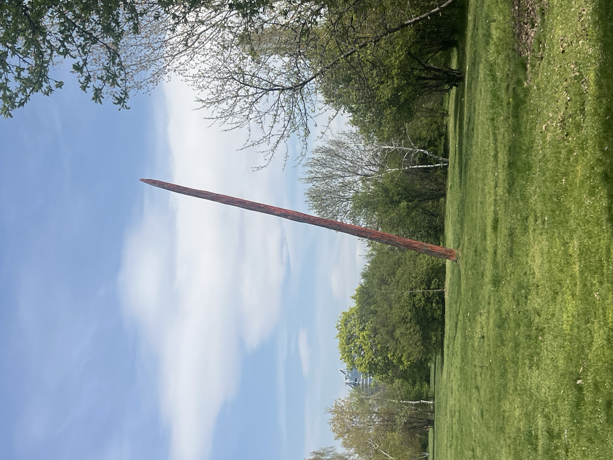 A vertical view of a landscape. The scene captures a green lawn, and trees under a blue sky with scattered clouds. A tall, possibly wooden structure stands prominently in the middle of the frame. In the background, a building can be seen. Based on the wooden structure and environment, it is likely to be a part of a sculpture or artwork. Without further information, it's impossible to ascertain the exact location.