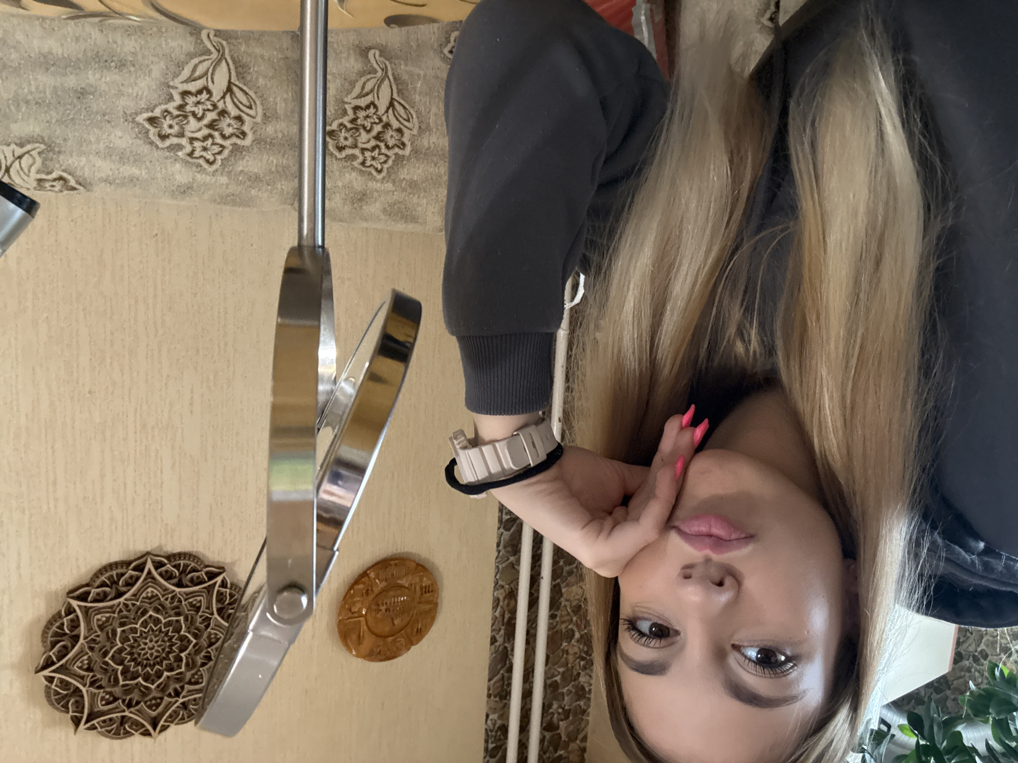 A woman is posing for a selfie. She has long blonde hair and is wearing a dark hooded sweatshirt and a white watch with a black strap. She is holding her face with one hand. Behind her is a light brown wall with a mirror, a mandala-style wooden wall decoration, and a decorative oval plaque. The picture was not taken in Sofia, Bulgaria.