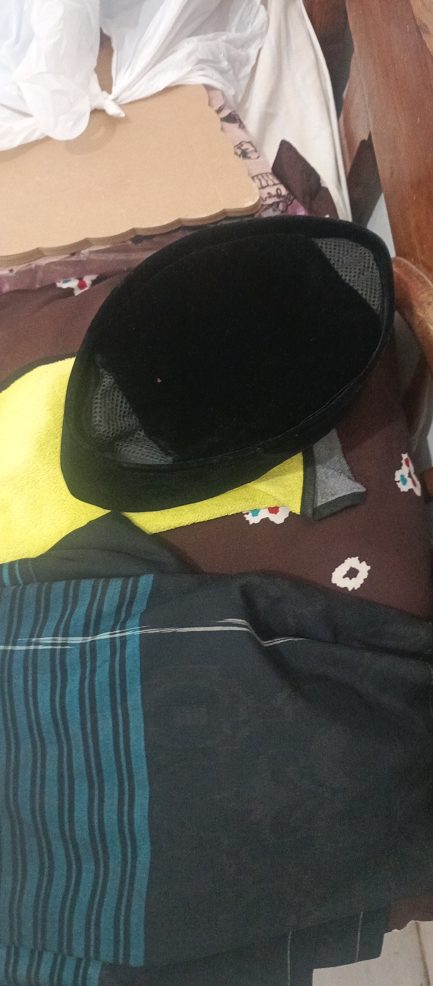 A black skullcap, likely a peci, rests on a yellow cloth atop a patterned brown fabric. To the right of the skullcap is a wooden surface, possibly a chair. Below the skullcap and to the left is a dark fabric with teal and black vertical stripes. The overall scene appears to be indoors, in a domestic setting within Arjasa, Indonesia.