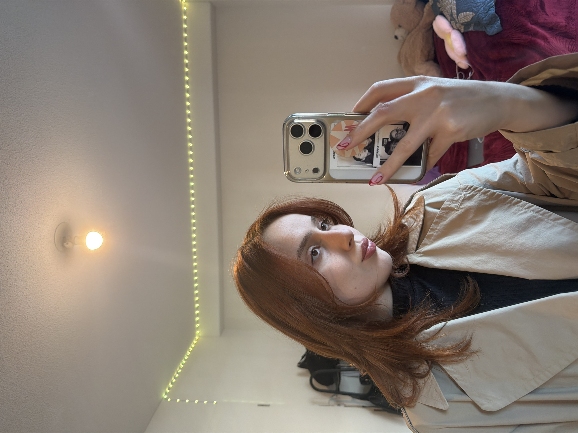 A young female with reddish-brown hair is depicted from the chest up, taking a mirror selfie in an indoor environment located in Strasbourg, France. She is wearing a black turtleneck under a beige trench coat. Her right hand, with red and pink abstract nail art, holds a smartphone with a clear case and an orange/white patterned grip. Small photos are visible within the phone case. The subject is looking to her right, with slightly pursed lips. The background reveals a room with white walls and a white ceiling featuring a single exposed light bulb and a string of green LED lights along the wall's perimeter. To the left, a dark object resembling a coat rack with a black bag is visible. To the right, a bed covered in a deep red textured blanket holds a light brown teddy bear and a small pink object.