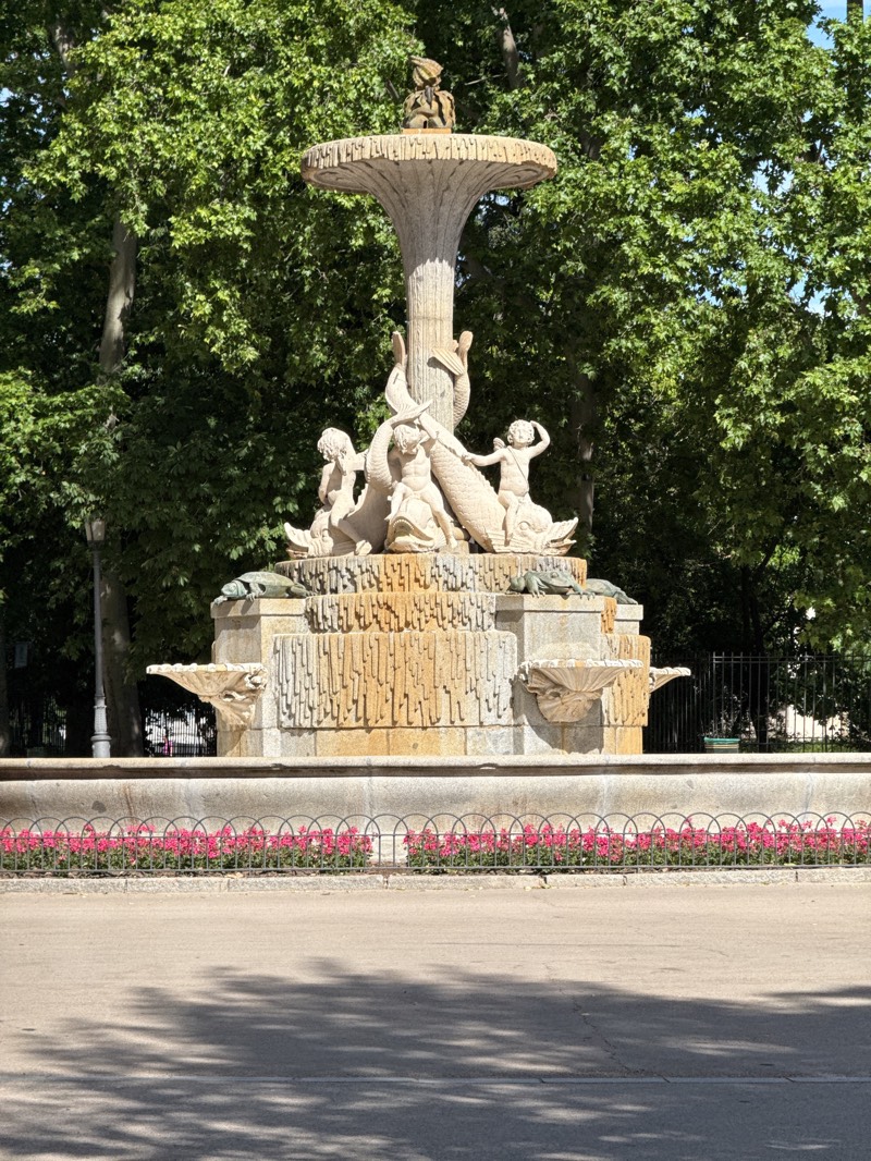 A large, ornate stone fountain is the central subject of the image, set within a lush green park. The fountain is crafted from light-colored stone and features multiple sculpted tiers.
The topmost tier is a mushroom-shaped cap, crowned by a small, solitary figure, possibly a putto or cherub. Below this, the middle section of the fountain showcases several cherubic figures actively interacting with large, stylized fish or marine creatures, their bodies intertwined in a dynamic composition. These figures appear to be playing or wrestling with the aquatic animals. The lowest tier consists of a rectangular base with a textured surface and smaller, shell-like basins protruding from its sides.
Behind the fountain, a dense canopy of vibrant green trees forms a natural backdrop, indicating a mature park or garden environment. In the foreground, a paved pathway or road runs across the bottom of the frame, marked by dappled shadows, suggesting strong sunlight. A low, decorative black metal fence separates the pathway from the fountain, and behind this fence, a neat row of bright pink flowers adds a splash of color to the scene.
The image captures a bright, sunny day, evidenced by the clear light and distinct shadows. The scene portrays a tranquil and aesthetically rich public space, characteristic of a grand urban park, consistent with the provided location context of Madrid, Spain. No specific activity or visible text is present in the image.