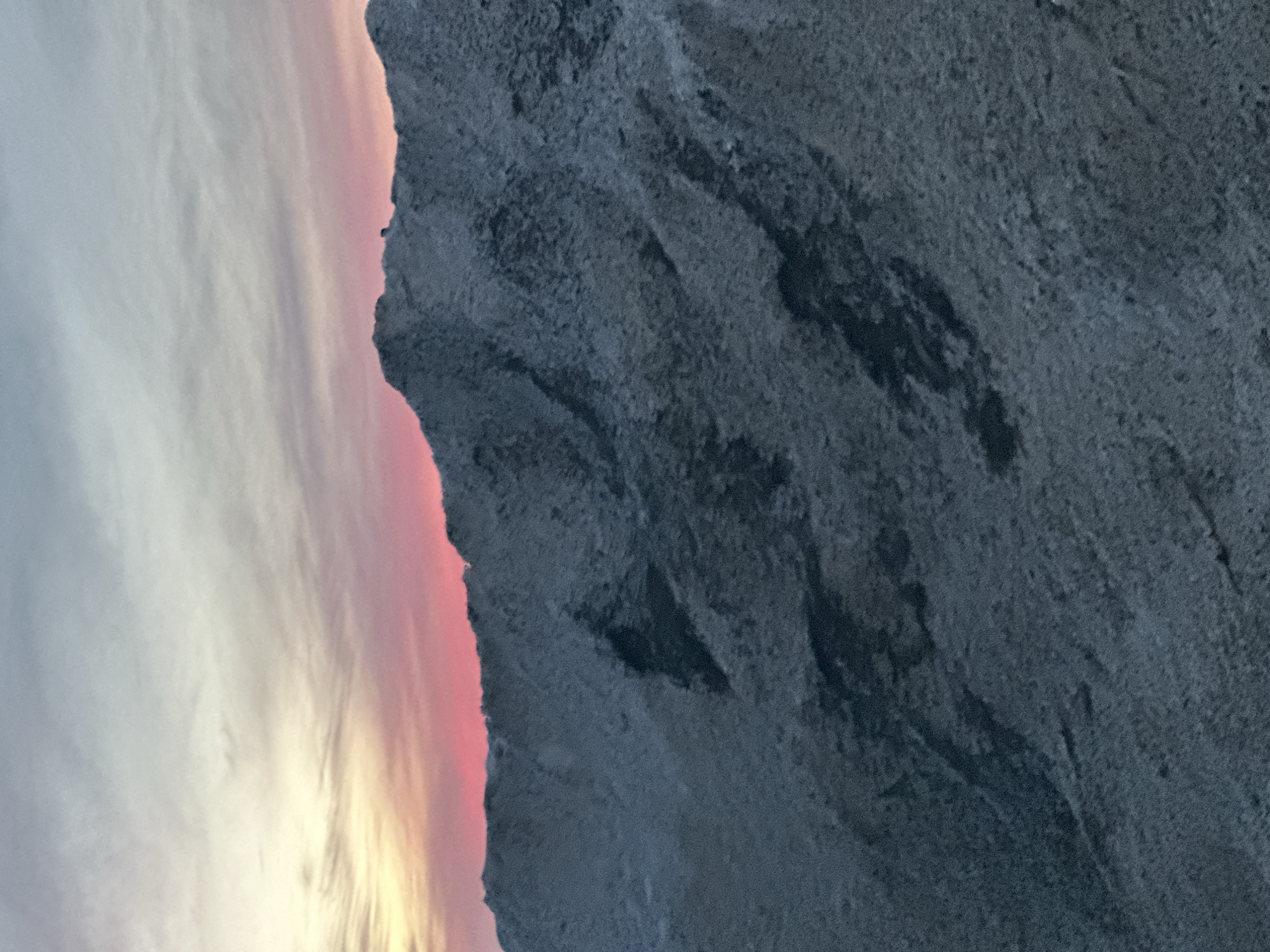 A panoramic view of a snow-covered mountain range, located in the city of Votkinsk, Russia, captured during either sunrise or sunset. The jagged peaks and slopes of the mountains, dark grey in tone, are dusted with a fine layer of white snow or frost, creating a textured, rugged surface. The sky above presents a striking gradient, transitioning from a light grey-blue at the top, to soft yellow hues, and culminating in a vibrant band of deep pink and red directly above the mountain crests. No human figures, structures, or discernible activity are visible within the frame. The overall environment suggests a cold, desolate high-altitude or winter landscape.