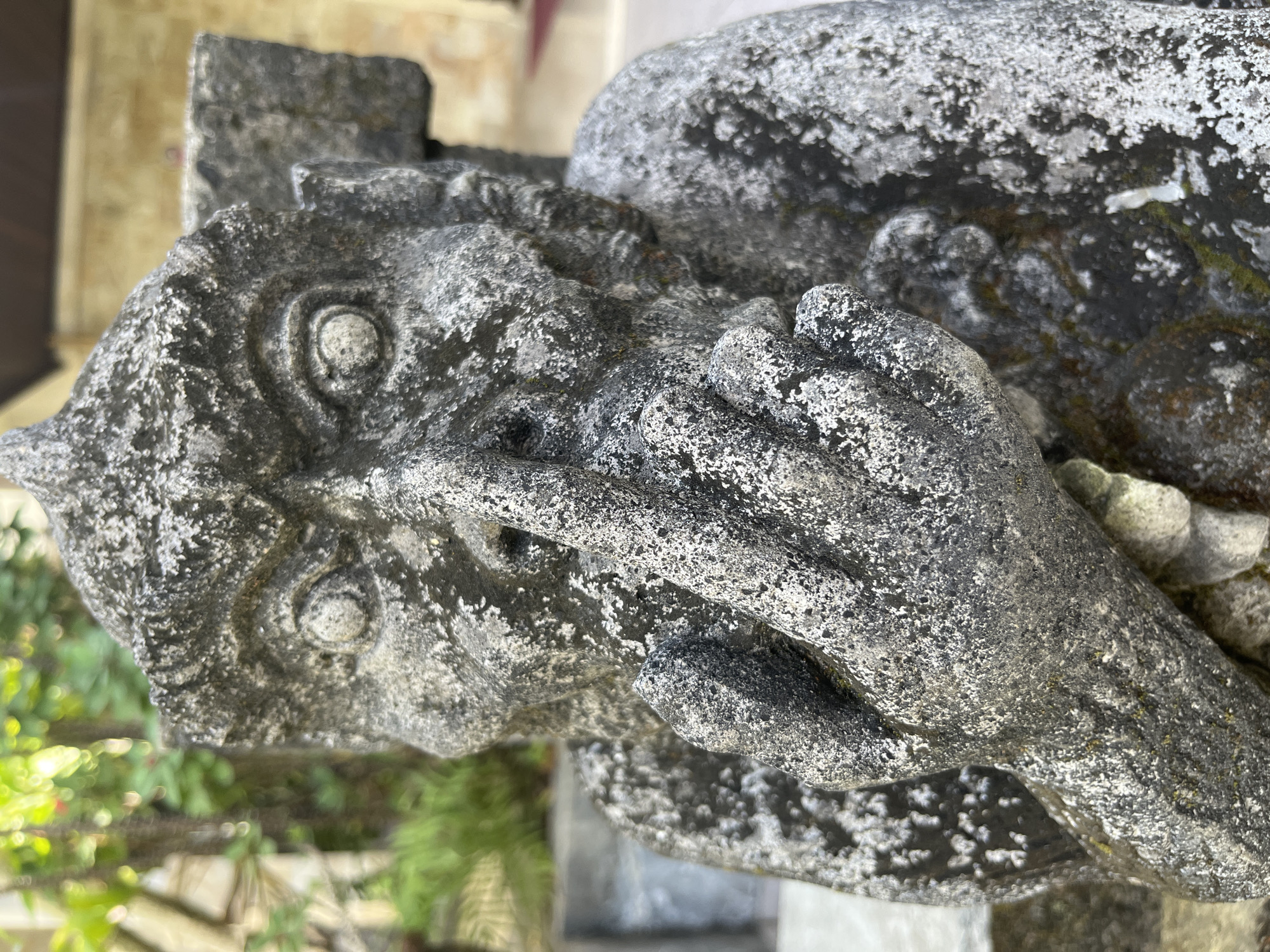 A weathered stone statue of a monkey with its finger to its lips is shown in a close-up shot. The statue is textured with moss and lichen, giving it an aged appearance. The monkey has large, expressive eyes and a stern facial expression. Its right hand is raised to its mouth, with the index finger gently pressing against its lips, in a gesture of silence. The statue appears to be outdoors, with blurred greenery and a hint of a building in the background, suggesting a garden or temple setting. The lighting indicates it is daytime, with sunlight casting shadows. The statue's posture and the gesture it makes are the central focus of the image. This statue is located in the Monkey Forest Temple, also known as Uluwatu Temple, in Kuta Selatan, Bali, Indonesia.