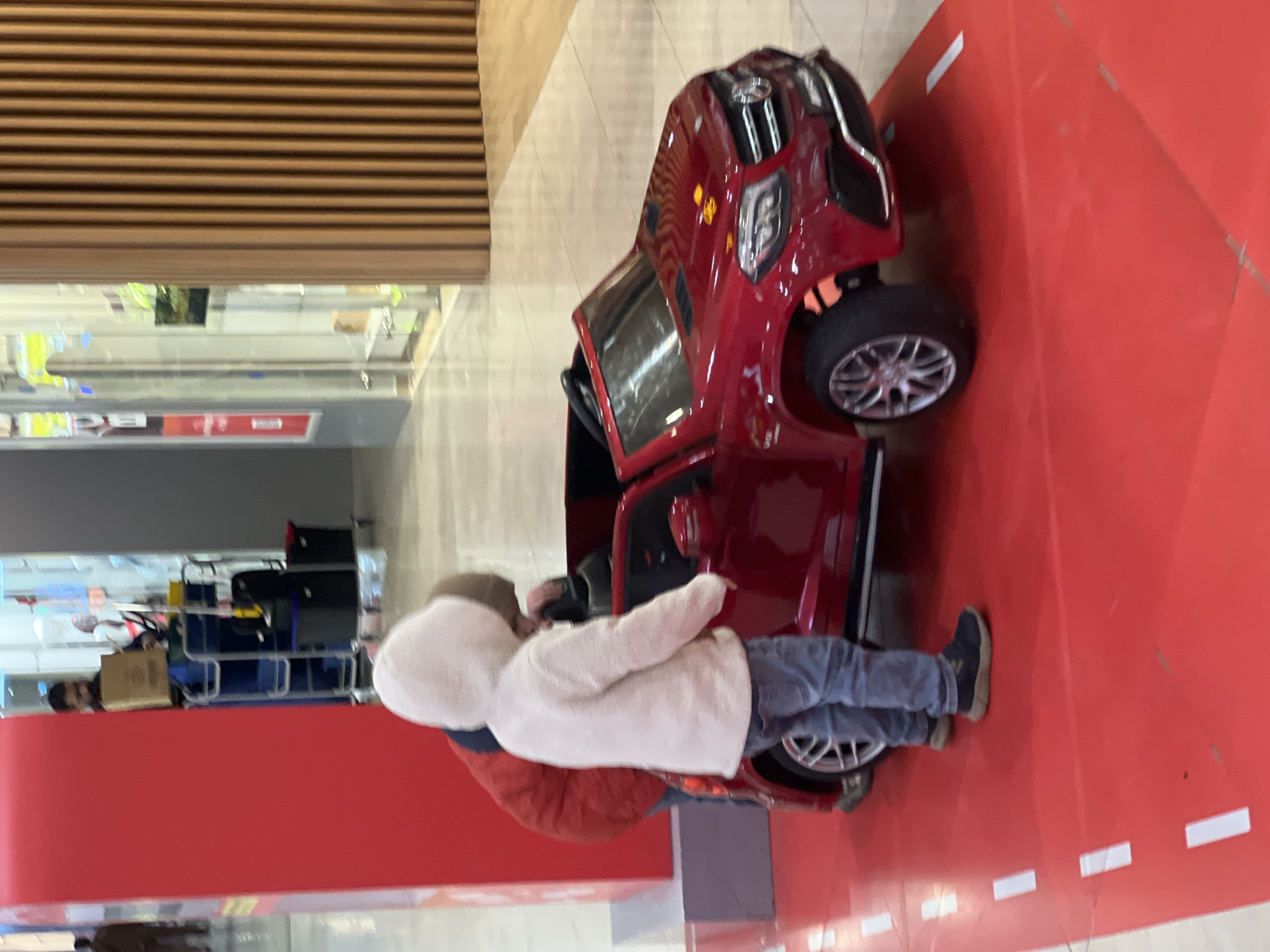 A child, clad in a hooded white jacket and blue jeans, is next to a small, red toy car inside what appears to be a shopping mall in Moscow, Russia. The child is near the open door of the toy car. The floor beneath the car is a bright red. The mall's interior features wooden slat paneling and large windows. A luggage rack filled with luggage is visible in the background.