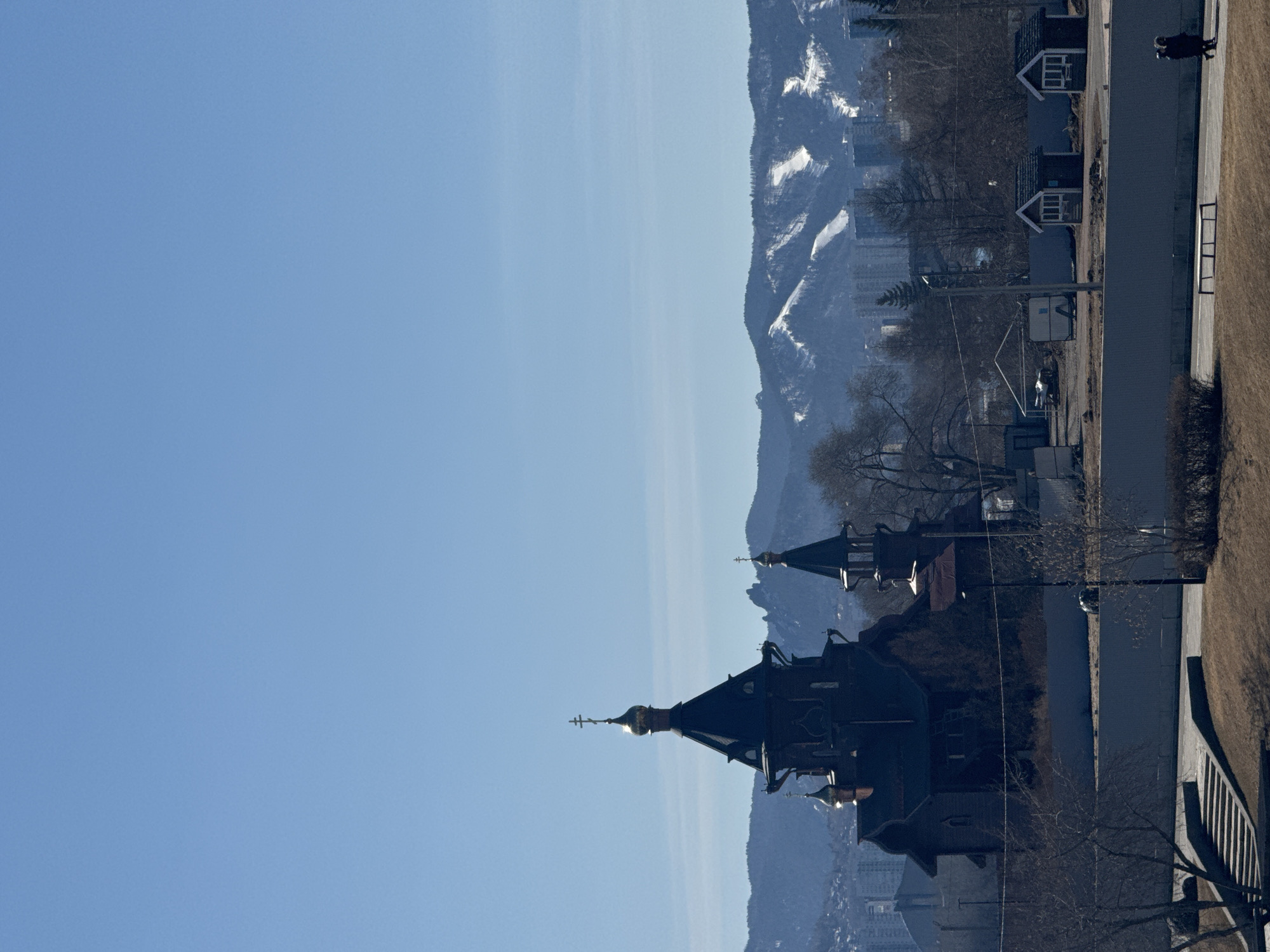 The image shows a partial view of the Paraskeva Pyatnitsa Chapel in Krasnoyarsk, Russia, set against a backdrop of distant mountains. The dark silhouette of the chapel’s spires with golden crosses is prominent in the foreground. A small road is visible to the right, with a brick wall and a grassy area, along with some small buildings, a light pole, and a few bare trees. In the upper right, a person is walking along the sidewalk. The sky is clear and blue, providing a stark contrast to the structures and the mountains behind. The mountain range appears to have patches of snow.