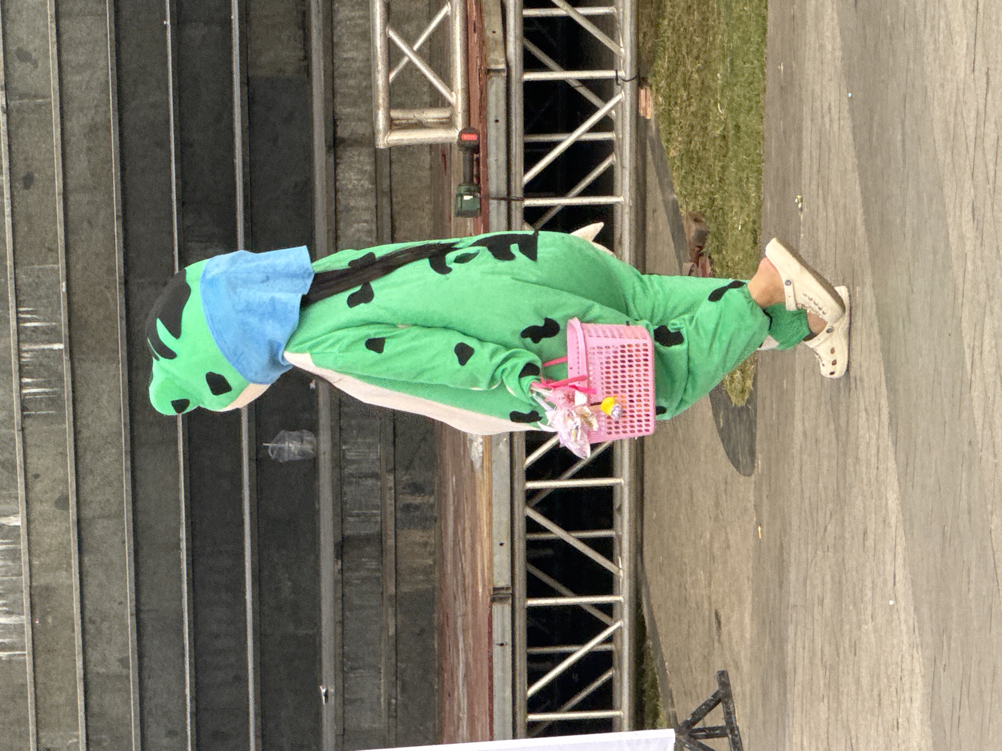 The image depicts a person walking from left to right, wearing a green and black spotted full-body costume resembling a frog, complete with a blue head covering. The person is carrying a pink woven basket filled with various items, including what appear to be candies or small toys. The scene takes place outdoors on a paved walkway in front of a tiered concrete structure that resembles stadium seating. In the background, there is a wooden platform with metal scaffolding underneath, suggesting a stage or an elevated area. A power tool with a red battery pack is visible on the platform. The ground is a mix of paved surfaces and grassy areas. The overall impression is of an event or performance taking place in an open-air venue. While the prompt specifies Da Lat, Vietnam, this information cannot be visually confirmed from the image alone.