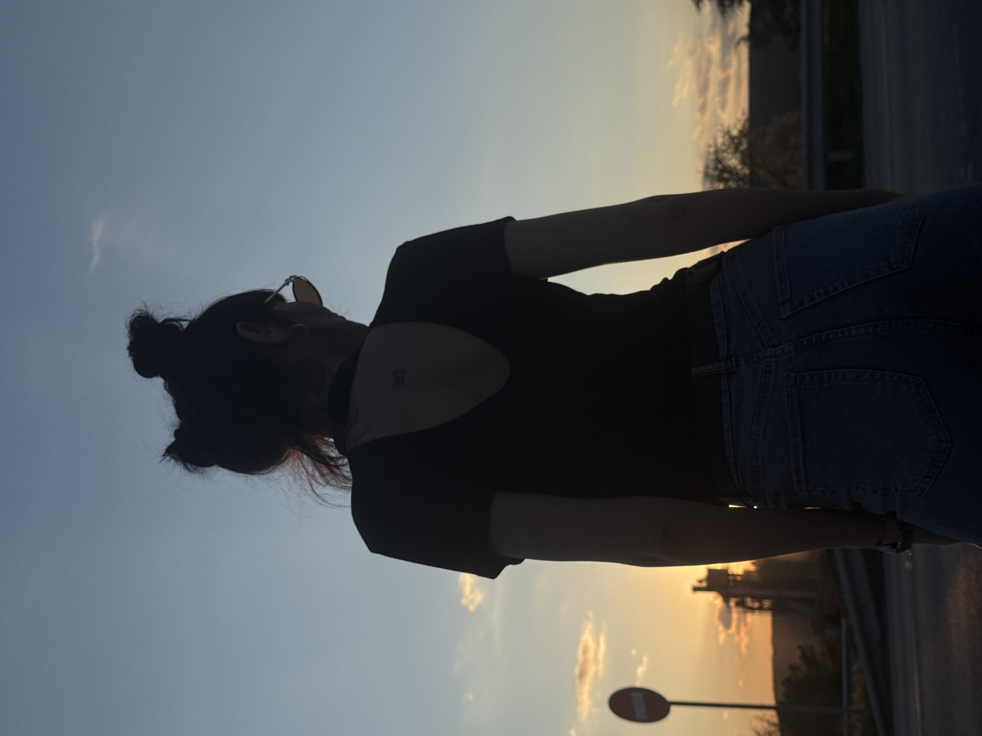 A woman stands silhouetted against a sunset sky. She has her hair in a bun, wears sunglasses, a dark top with an open back, and denim jeans. The background reveals a roadside with infrastructure, including a no entry road sign and foliage, suggestive of a location in or near the city of Kipra, Bulgaria.
