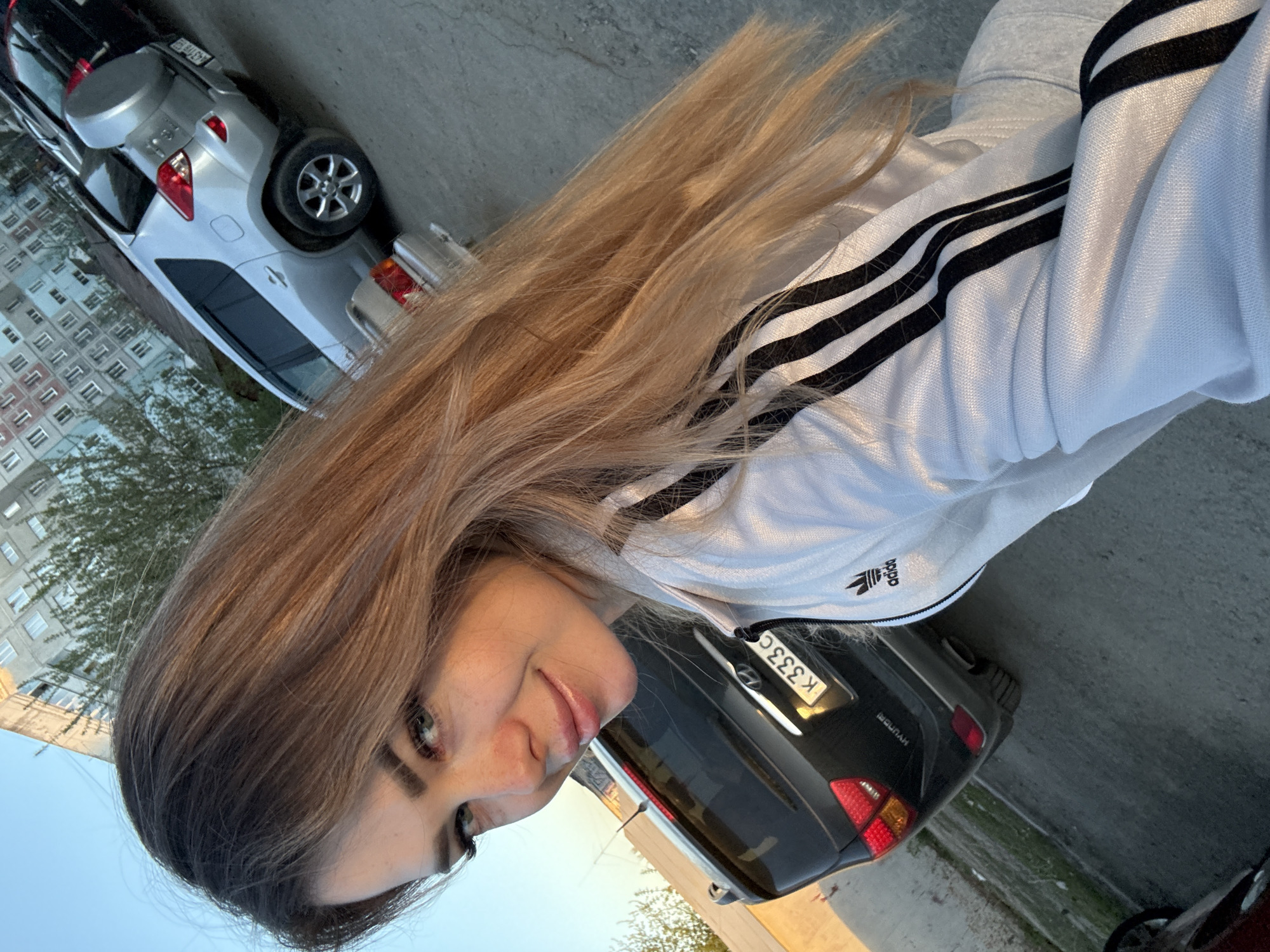 A woman with light brown hair and fair skin is standing outside in an urban area. She is wearing a white Adidas jacket with black stripes. Parked behind her is a black car with a license plate beginning with "K333C". In the background, there is a residential building and a silver car. The environment suggests an everyday street scene, with the presence of cars, a building, and pavement.