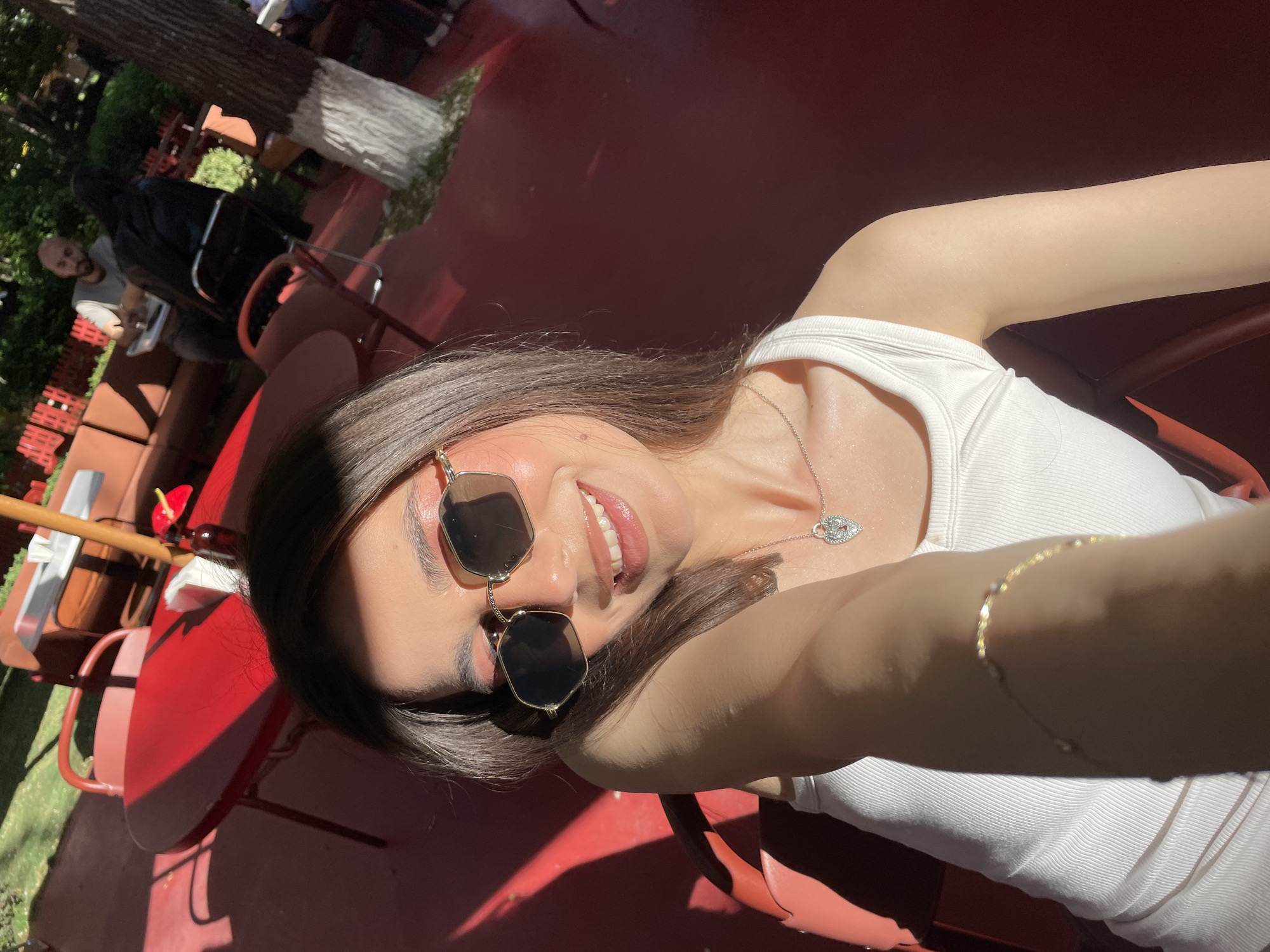 The image depicts a person with brown hair and fair skin, wearing a white tank top and gold-framed sunglasses, smiling and taking a selfie outdoors. The location appears to be a patio or outdoor seating area, evidenced by the red-painted surfaces, tables, and chairs. In the background, there is a tree with a whitewashed base and glimpses of other people seated. The scene is brightly lit, suggesting a sunny day. Without additional location context, it is impossible to confidently assert the location within Yerevan, Armenia.