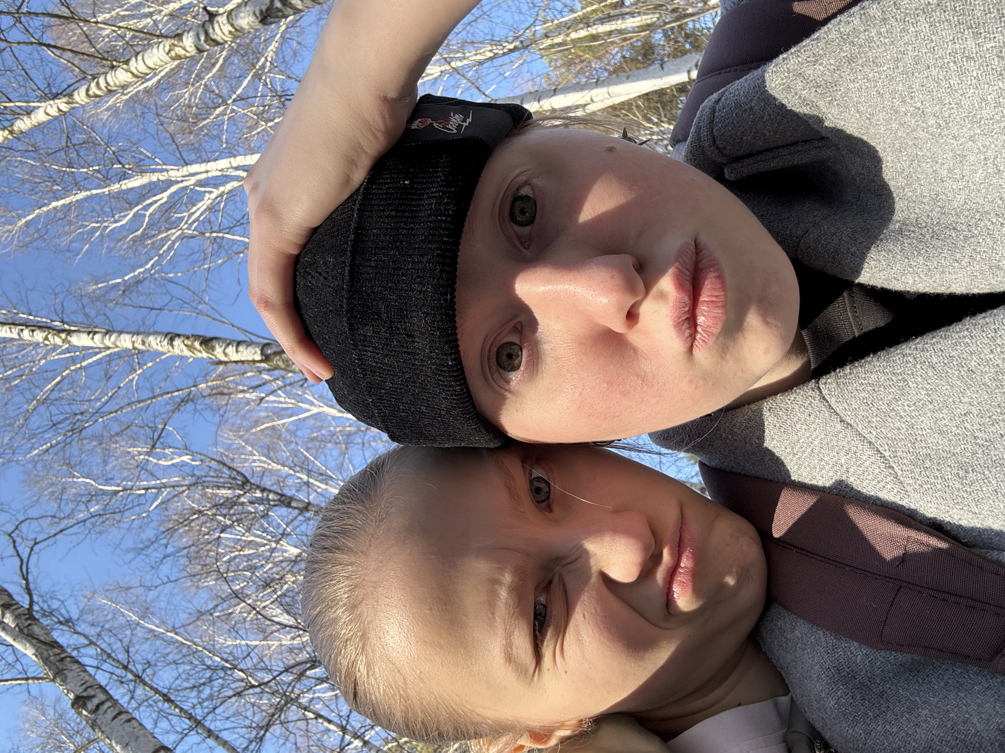 Two light-skinned individuals are visible in an outdoor setting, presumably in Perm, Russia. The upper person wears a black beanie and a gray coat or jacket, and is holding their hand near their head. The lower person's head is positioned near the upper person's shoulder. Bare trees are visible in the background against a light blue sky.