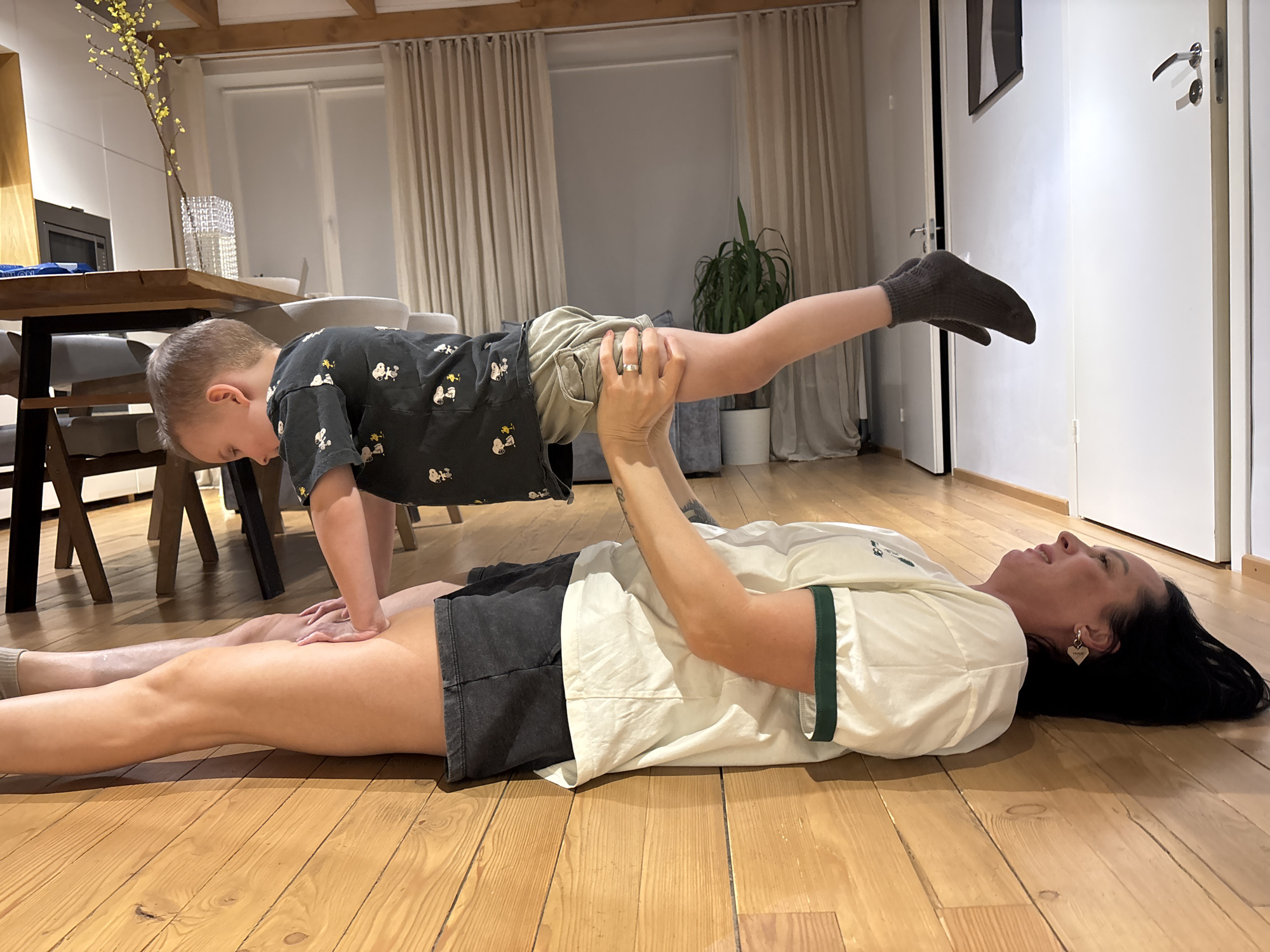 A woman is lying on her back on a wooden floor, holding up a young child in a plank position. The woman is wearing a white t-shirt with green trim, dark shorts, and has dark hair. The child is wearing a dark patterned shirt, tan shorts, and socks. The scene is indoors, with a table and chairs visible in the background, along with a window and curtains. Based on information available, the location within Chaikovsky, Russia, cannot be determined.