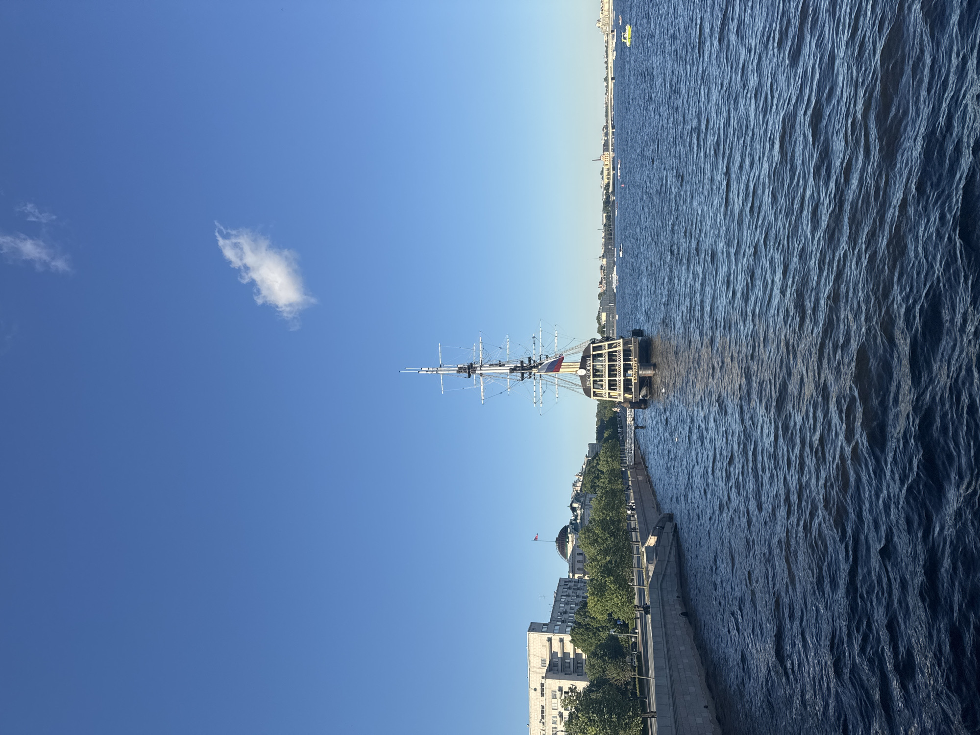 This is a photograph taken from a bridge over a body of water, likely a river or canal, in Saint Petersburg, Russia. The water is choppy and a deep blue color. In the center of the frame, a large, dark sailing ship with multiple masts is docked along the embankment. The ship has the word "РЕСТОРАН" (Restaurant) written on its side. Along the embankment, there is a paved walkway lined with trees. Behind the trees, several multi-story buildings and a domed structure are visible. The sky is a clear, bright blue with a single, small white cloud. Several smaller boats and a yellow watercraft are present on the water in the distance.