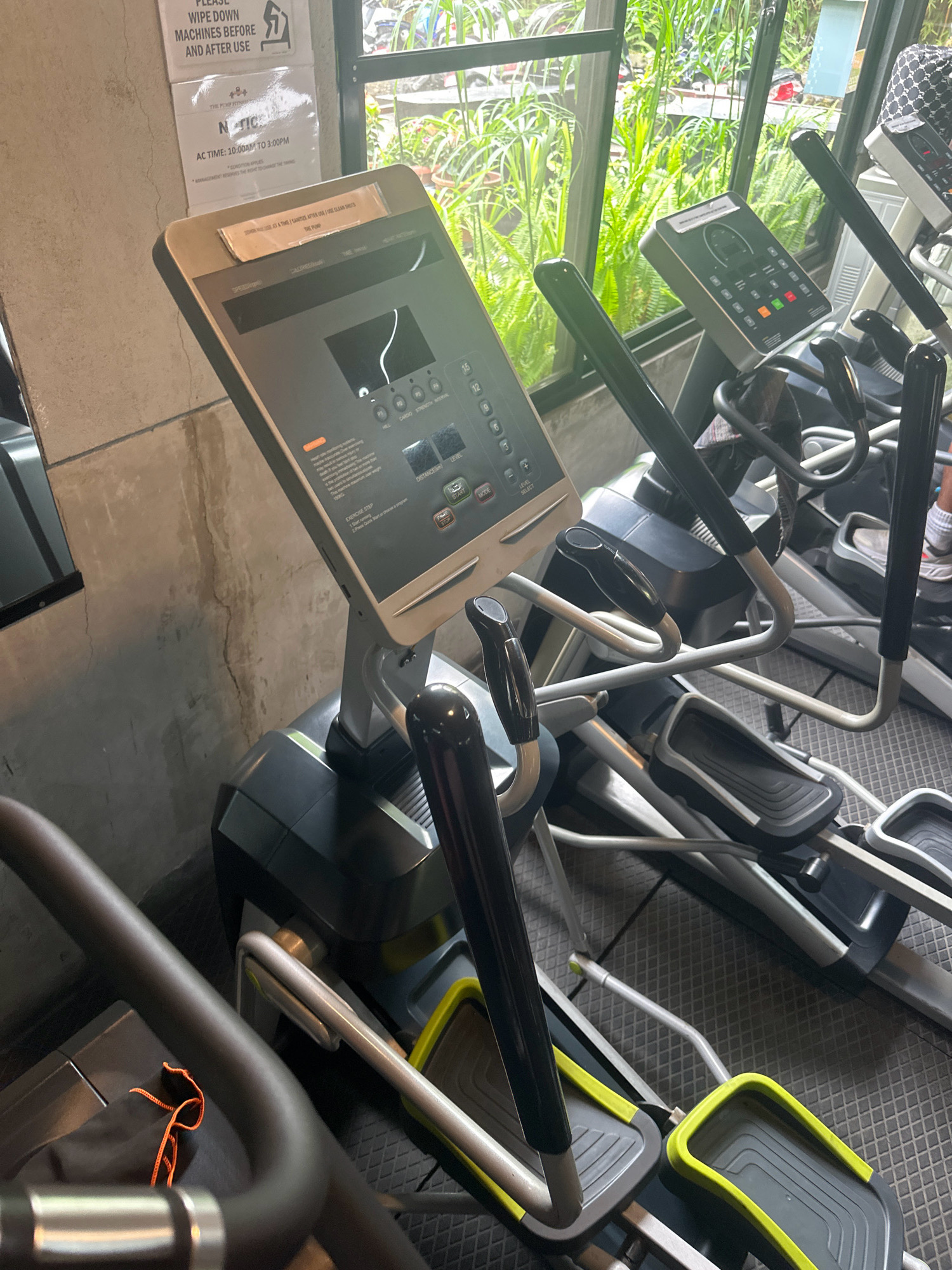 In Lalitpur, Nepal, within a gym setting, an indoor elliptical machine is prominently displayed in the foreground, angled slightly to the right. Its control panel, featuring various buttons and a digital display, is visible. The machine is set against a textured wall on the left and a large window with lush greenery and foliage visible through it on the right. Other exercise equipment, including another elliptical machine and parts of what appears to be a treadmill, are present in the background. The floor is covered with a dark, textured matting. A sign is affixed to the wall above the machine, containing instructional text related to machine usage and gym hours.
