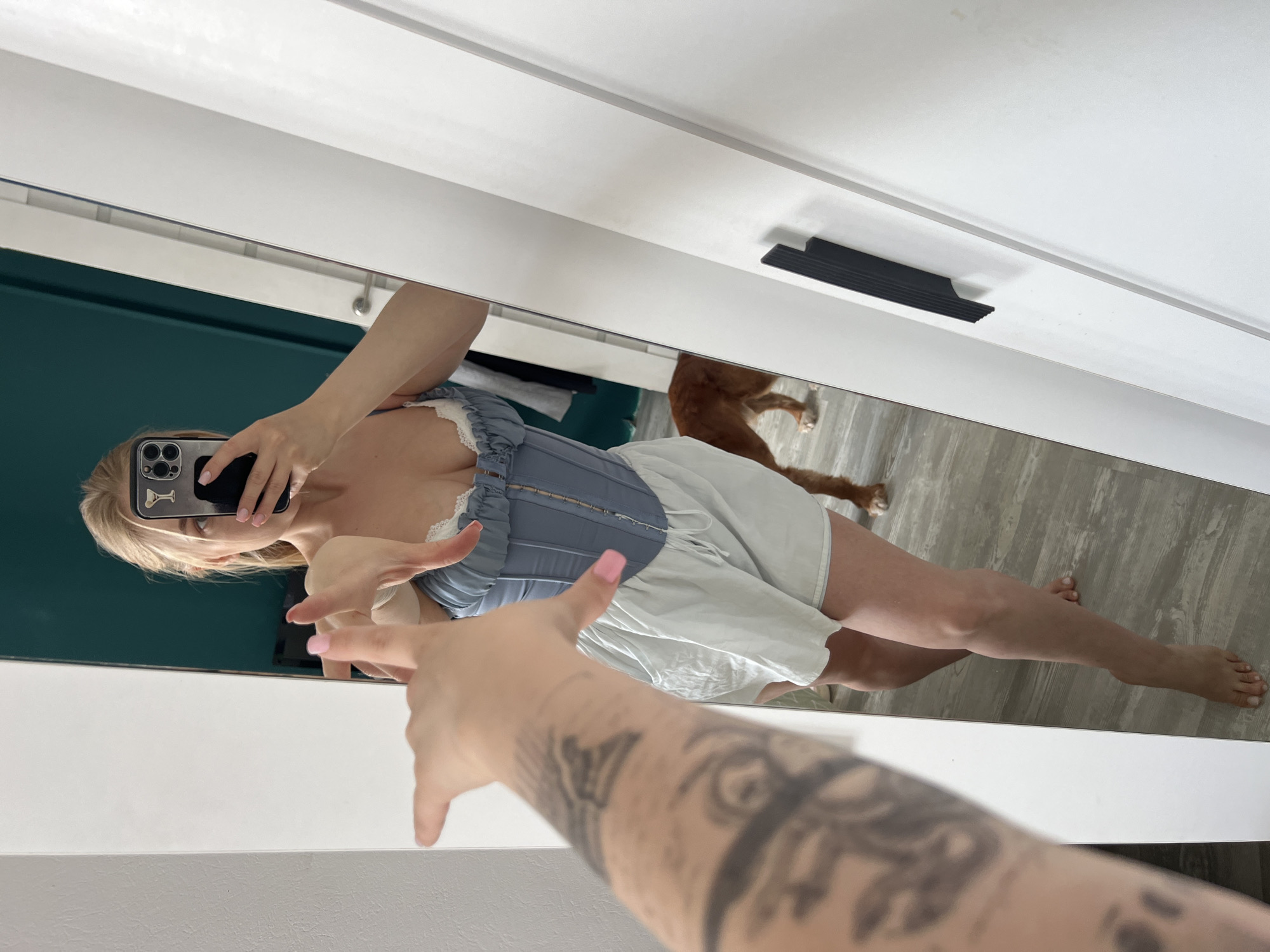 A full-length mirror reflects a woman posing in what appears to be a bedroom. She is wearing a light blue corset-style top with white lace trim and a pair of light-colored shorts. Her blonde hair is pulled back, and she is holding a smartphone in her right hand to take the picture. Her left arm is extended forward, and her hand is positioned as if reaching out. In the background, a medium-sized, reddish-brown dog stands near the wall. The floor appears to be laminate wood. The mirror is framed by white closet doors, one of which has a black handle. The image does not provide any specific details that would allow for its location to be identified as the city of Tyumen, Russia.