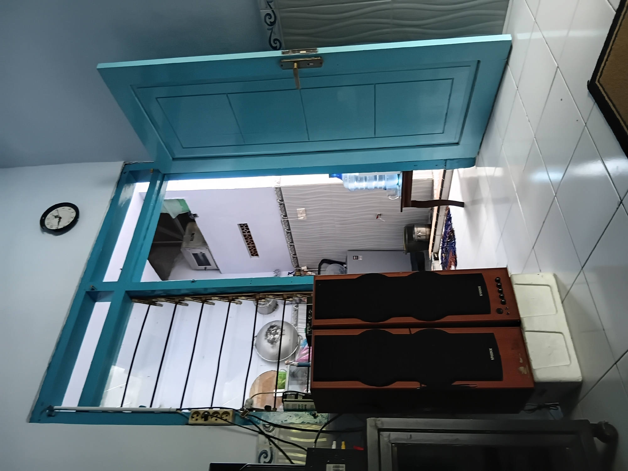 The image captures a sunlit interior scene, likely in Tanggul Wetan, Indonesia, focusing on a doorway and its surroundings. A bright blue, paneled door stands open, revealing a glimpse of an adjacent room or hallway. To the left of the doorway, a louvred window with horizontal slats allows natural light to enter, illuminating a wall-mounted electrical outlet and various cables.
Dominating the foreground are two tall, brown wooden speakers with black grilles, positioned on top of a white rectangular object. A metal cabinet with a glass door and wheels is partially visible to the left of the speakers. Beyond the speakers, through the open doorway, a kitchen-like area is visible, featuring a large water dispenser, a small wooden table, and kitchen utensils like pots and pans hanging on a rack. A silver refrigerator is also present. The floor is tiled with large white squares. A round clock with a black frame and white face is mounted high on the wall near the ceiling. The overall impression is of a lived-in, functional space.
