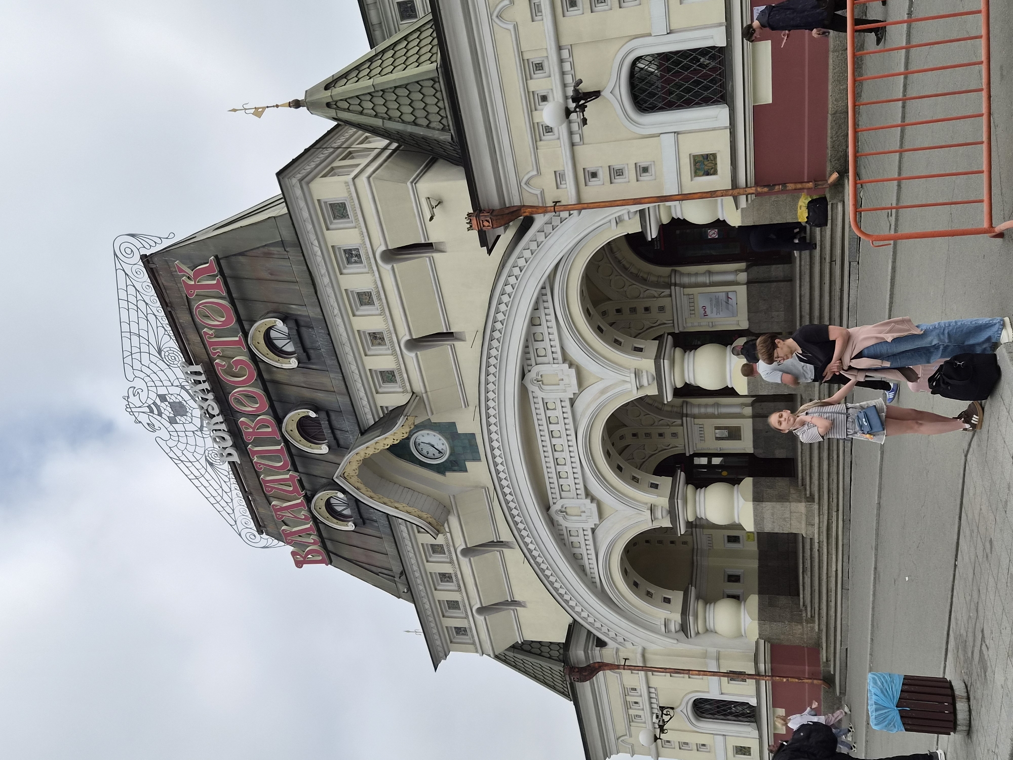 The image shows the facade of the Vladivostok railway station in Vladivostok, Russia. The station's name, "ВЛАДИВОСТОК", is prominently displayed in large red letters on the roof. The building features a grand architectural style with arches, columns, and decorative elements. The sky is overcast.
In the foreground, two young women stand facing the camera. The woman on the left is wearing a striped t-shirt, denim shorts, and a small blue crossbody bag. She has light brown hair and is looking at the camera. The woman on the right is wearing a black t-shirt, blue jeans, and a light pink cardigan draped over her shoulders. She is also holding a phone and has dark hair. Both women are wearing sneakers.
To the left of the women, a blue trash can with a blue plastic bag liner is visible. Further left, a lamppost is attached to the building. In the background, the entrance to the train station is visible with several people entering and exiting, some carrying luggage. An orange metal barrier runs across the lower right portion of the image. The ground in front of the station is paved, and there are steps leading up to the entrance.