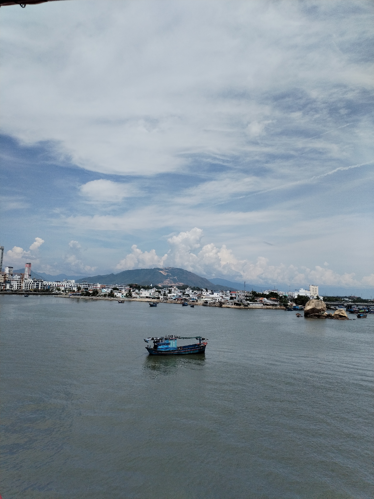 The image depicts a panoramic view of the city of Nha Trang, Vietnam, with a wide expanse of dark water in the foreground. A blue and dark-colored boat with a cabin is positioned in the middle of the water, with a rope extending from its stern. The background features a shoreline lined with buildings of varying heights and architectural styles, including some taller modern structures and lower-lying houses. Beyond the city, rolling hills and mountains form the backdrop, with a hazy, lighter-colored mountain range visible in the distance. The sky is partly cloudy, with patches of blue sky interspersed with white and gray clouds. Several other boats are scattered throughout the water closer to the shore. The scene is captured from a slightly elevated perspective, offering a broad view of the coastal urban landscape.