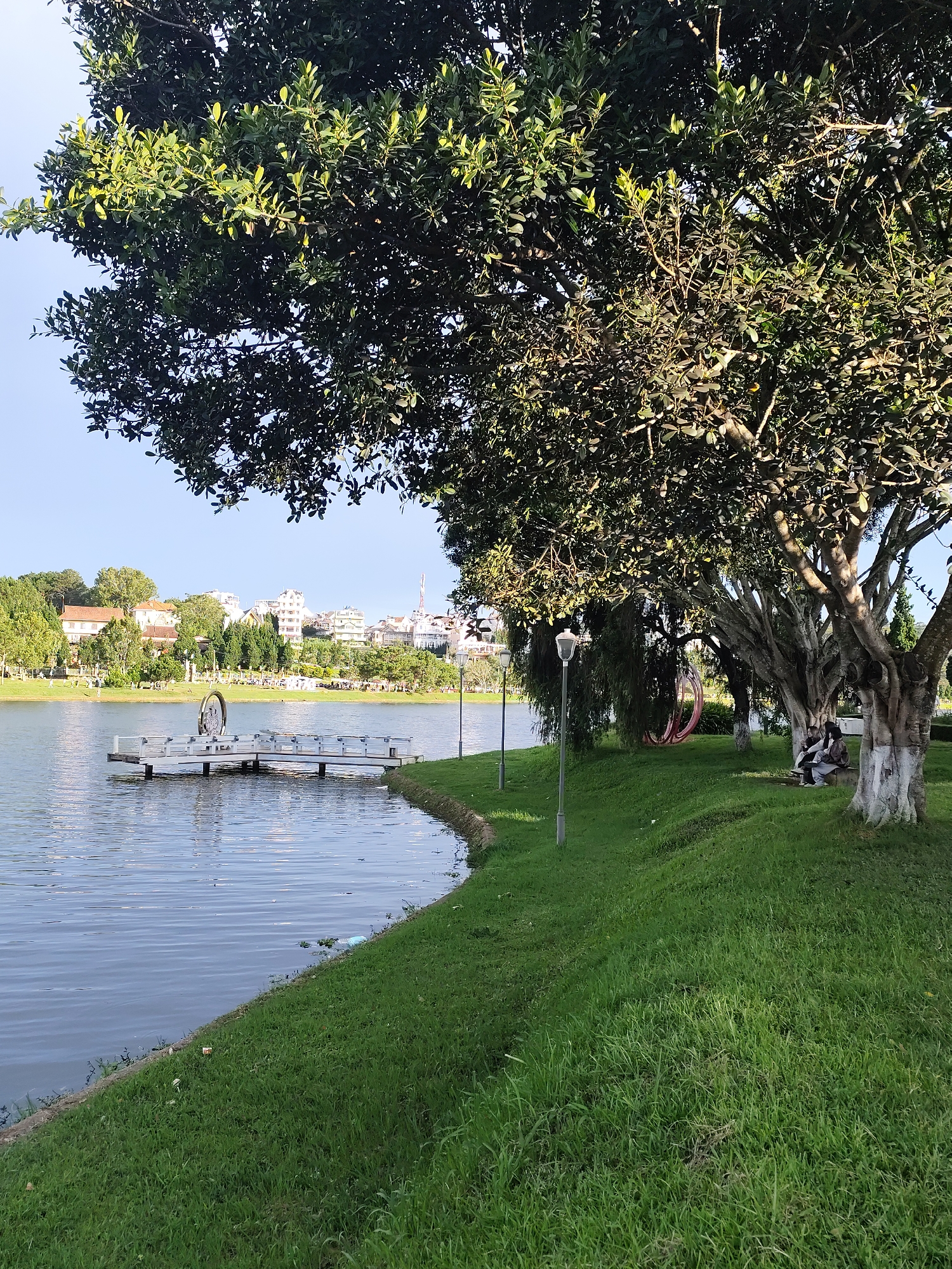 The image shows a scenic view of Xuan Huong Lake in Dalat, Vietnam. A white floating dock with several benches is anchored in the calm, rippling water. The foreground features a lush green lawn sloping towards the lake, with several large trees providing shade. In the mid-ground, a tall lamppost stands near the edge of the water. Further in the distance, across the lake, a cityscape is visible with buildings nestled amongst trees. On the right side of the image, under the shade of a large tree, two people are seated on a bench, seemingly conversing. The sky is clear and blue, suggesting fair weather.