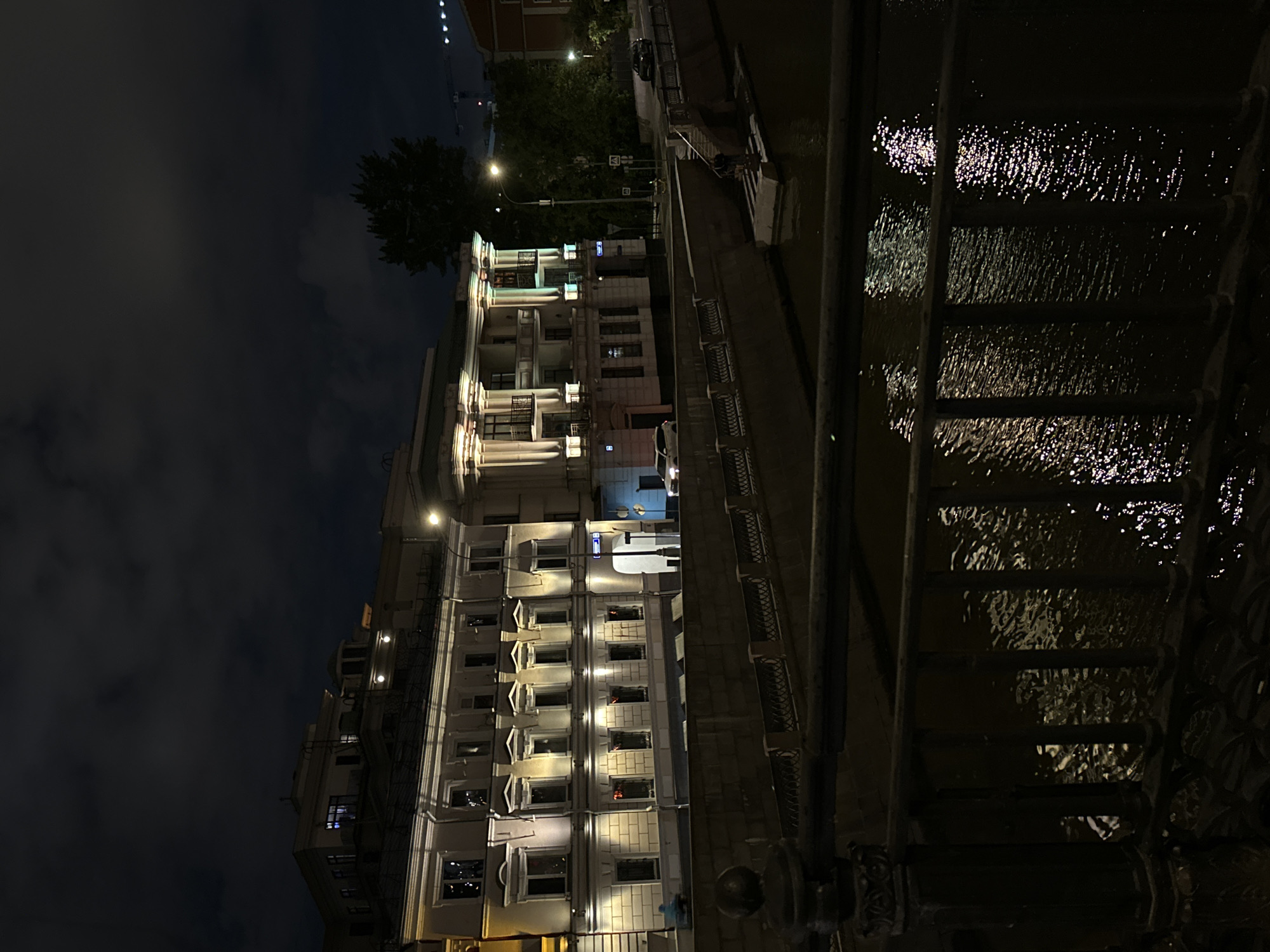 At night, the camera faces a long, lit building with many windows. To the right of the building, water reflects the lights. A dark sky looms above the building. A metal railing is visible in the lower right corner. The location is Unknown, Unknown.